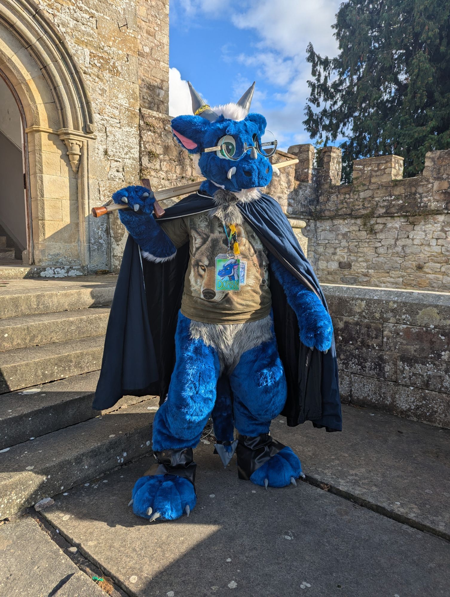 Blue dragon fursuiter Doran stands guard in front of a castle entry stairway, wearing a blue cloak and relaxing with a wooden longsword resting on his shoulder.