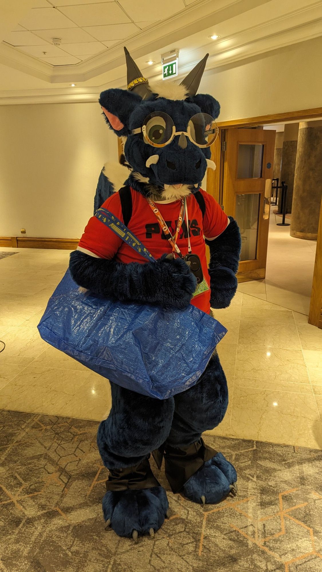 Blue dragon fursuiter Doran, standing in a hotel lobby with a blue IKEA shopping bag over his shoulder.