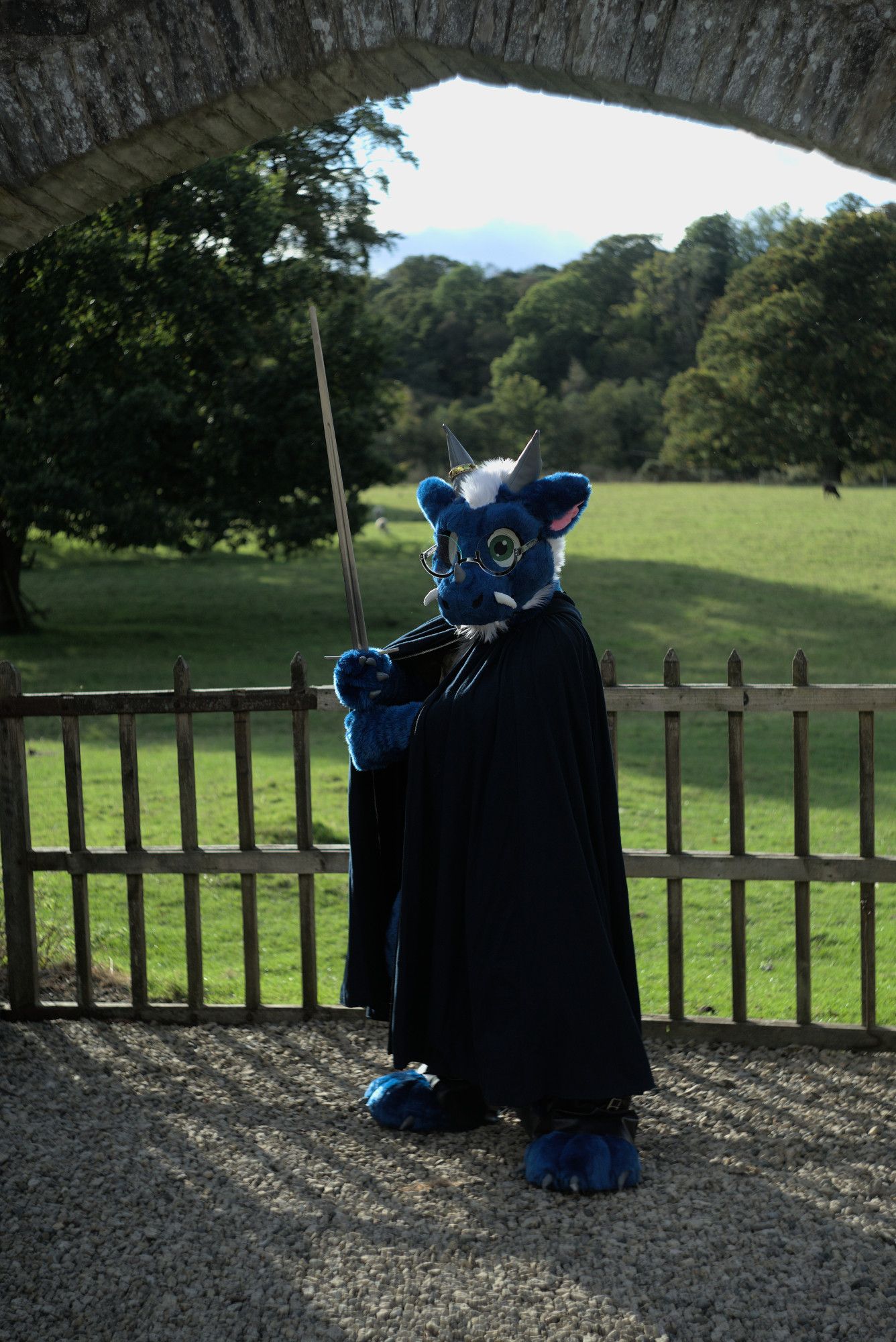 Blue dragon fursuiter Doran poses before a castle gate with a sunny green pasture in the background. He is wearing a dark blue cloak and holding a steel longsword in a vertical guard position, looking ready to battle.