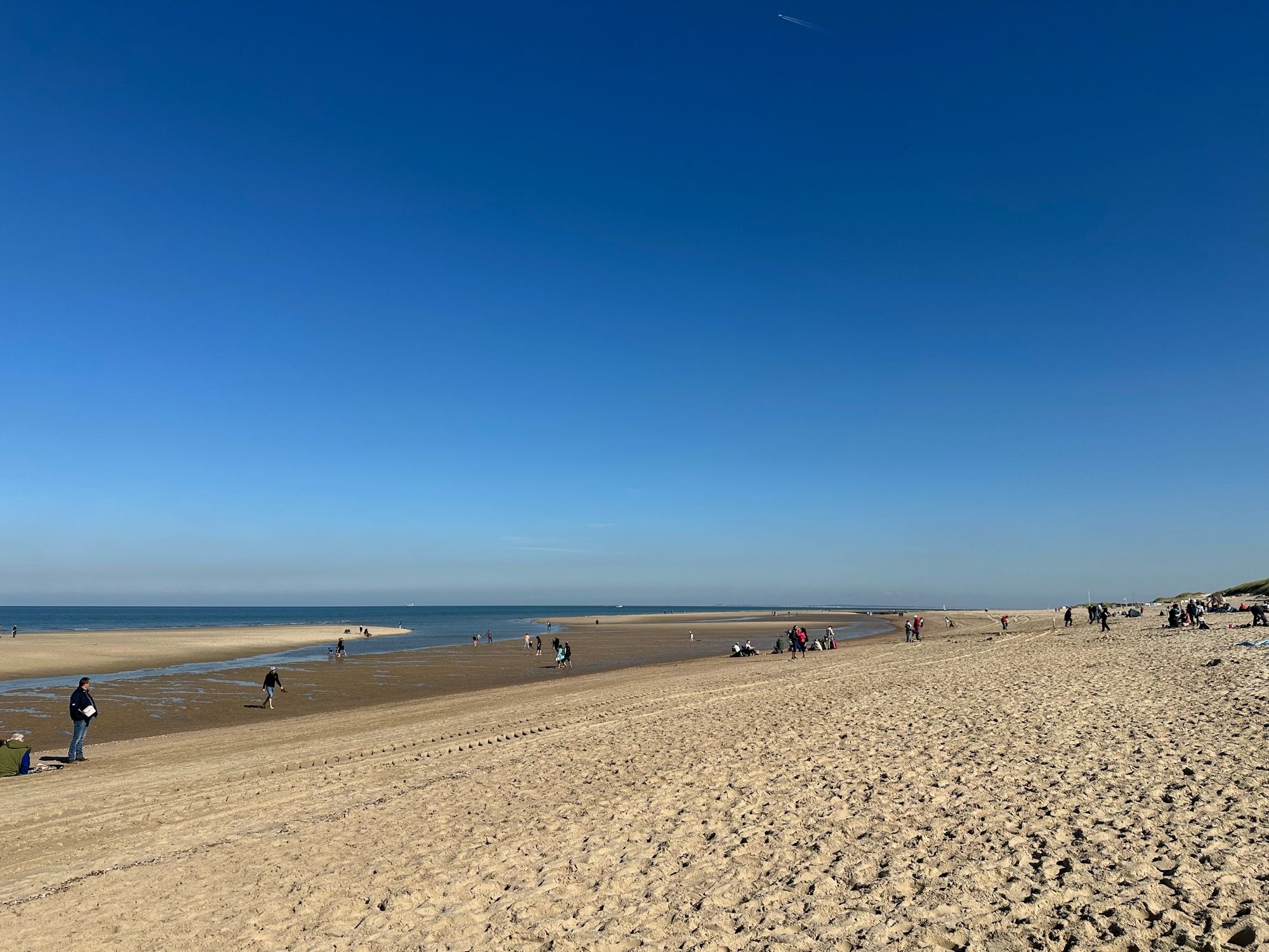 Blick vom Strand ans Meer. Vor dem Meer ist ein fast leerer Priel. Strahlend blauer Himmel.