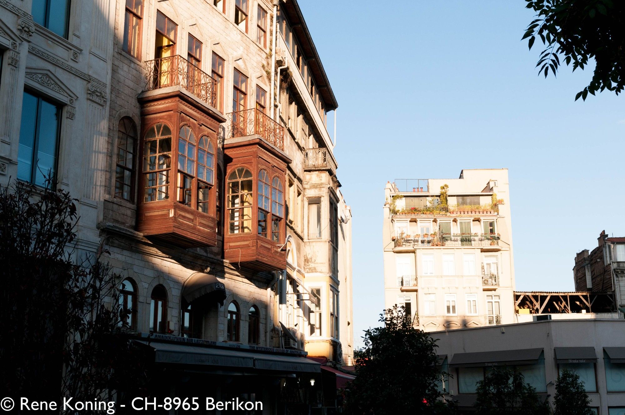 Eine Häuserzeile zieht sich von Links einer Strasse entlang, 2 Erkern scheuen heraus. Die Sonne scheint am Abend über die Fassade und wirft vorne einen Schatten auf dem Gebäude während die Erker noch in der Sonne stehen