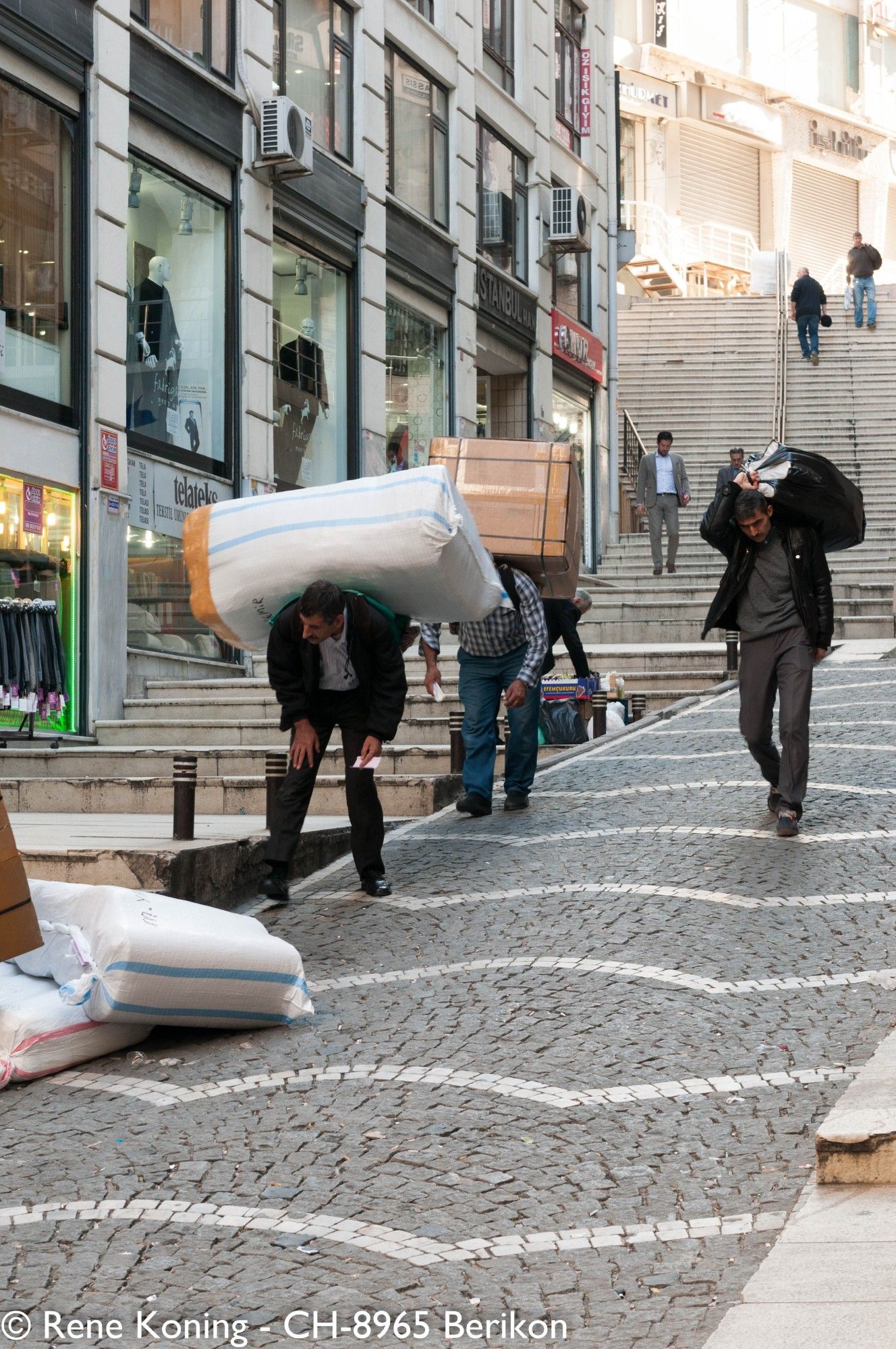 An einer steilen Strasse, im Hintergrund ist ein Häuserblock sichtbar, stehen 2 Männer krumm gebogen. Ihre Oberkörper sind uns zugewandt.  Auf Ihren Rücken haben beide jeweils eine sehr grosse und schwere Last. Diese Art von Warentransport ist in Istanbul noch sehr gängig. Rechts von den beiden Männer kommt ein Mann aufrecht gelaufen mit einem Sack auf dem Rücken, er lauft gerade. Im Bildhintergrund geht eine Treppe zwischen den Häusern hoch.