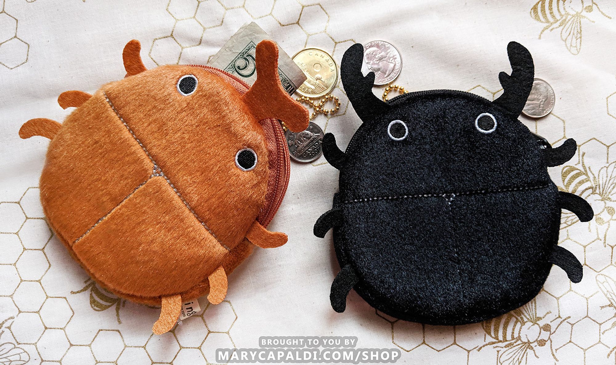 Photo of two round coin pouches made to look like beetles. Each has six legs made of felt, embroidered black eyes with white outline, and lines embroidered on its body to look like elytra. The brown one on the left has a single branched horn, while the black one on the right has two jaws, both made of felt. There is some money, mostly coins, scattered beneath them on a light-colored cloth patterned with honeycomb and honeybees.