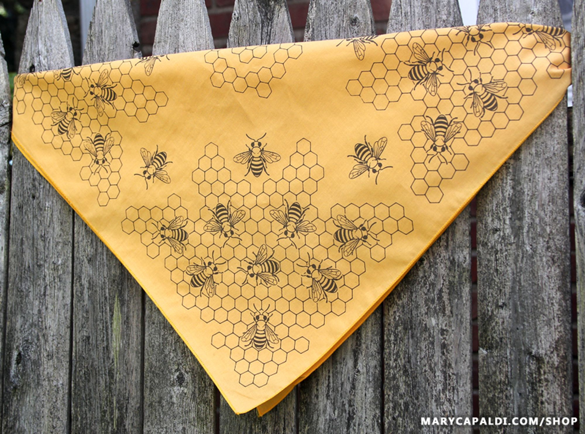 Photo of an athletic gold bandana with a honeybee and honeycomb design printed in warm brown ink. It's folded in half diagonally and resting on a worn wooden fence as a backdrop.