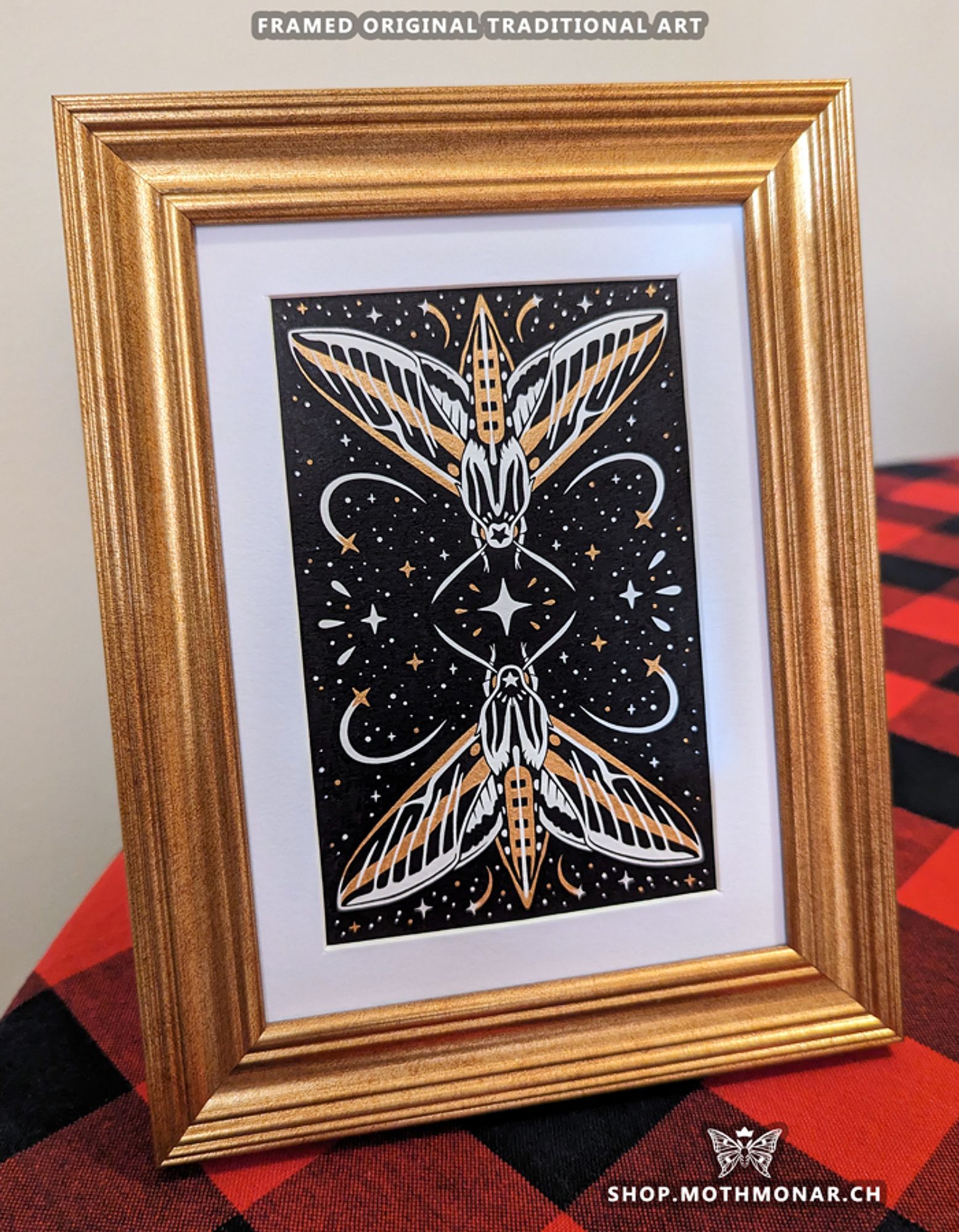 Framed traditional art piece in black, white, and gold featuring two white-lined sphinx moths. Each has a five-pointed star on their head and is reaching toward a large four-pointed star in the middle. Lots of other stars and accents surround them. The piece is matted in white and set in a gold frame sitting on a red plaid tablecloth.