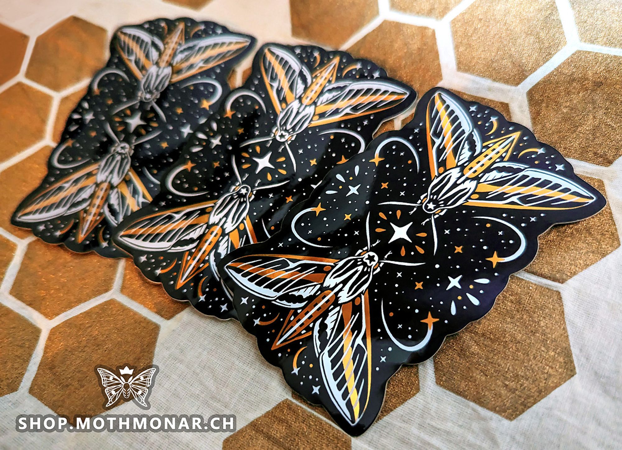 Photo of a spread of three identical die-cute vinyl stickers that are black, white, and shiny metallic gold featuring two white-lined sphinx moths. Each has a five-pointed star on their head and is reaching toward a large four-pointed star in the middle. Lots of other stars and accents surround them. The stickers rest on a cloth that is pale with an irregular pattern of solid muted gold hexagons. The watermark is http://shop.mothmonar.ch with a logo of a crowned moth.