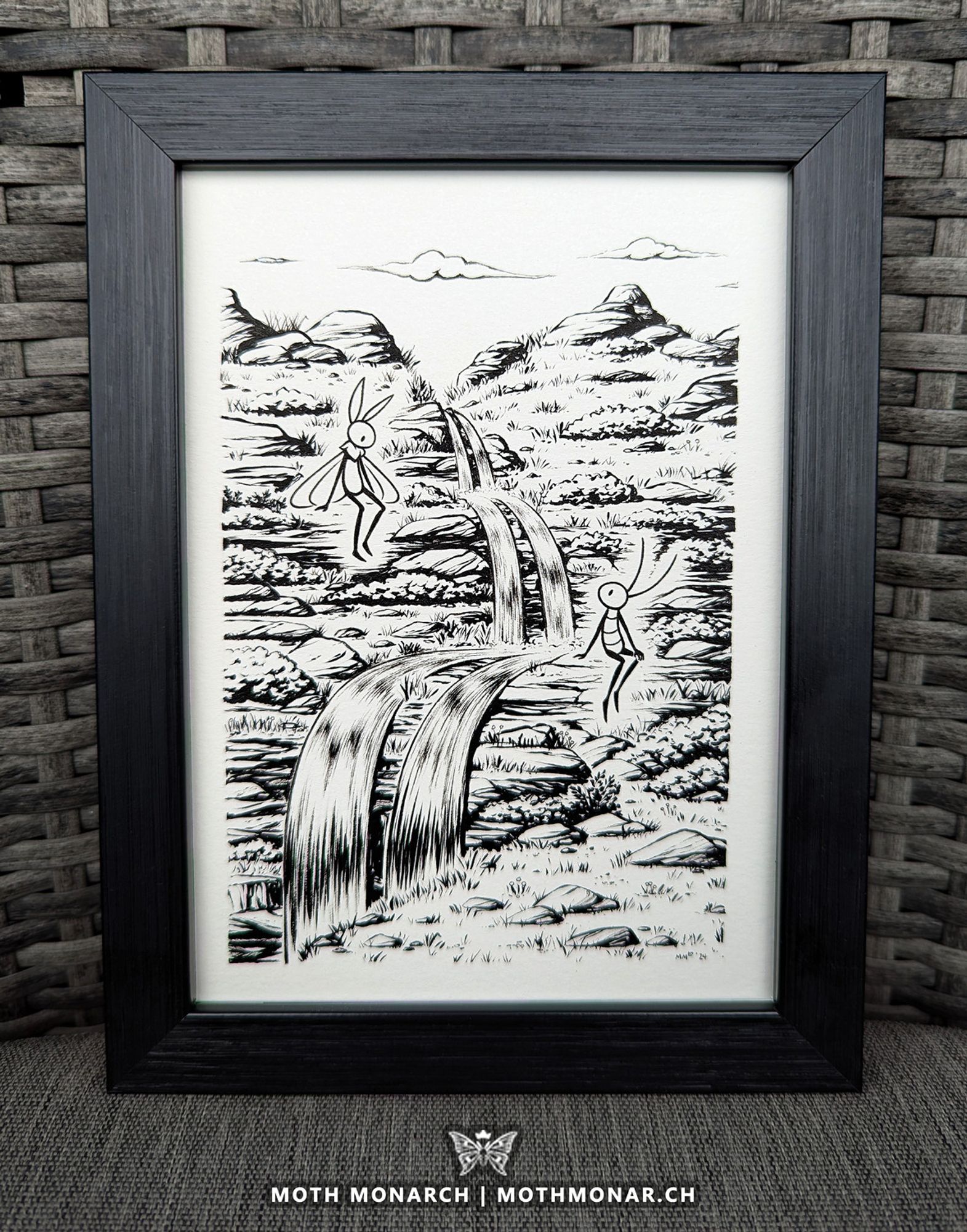 Photo of a framed artwork on a gray chair. It's a black and white ink piece with two cartoon insects sitting on either side of a split waterfall cascading in the middle of the image. They are meeting each other's gaze. The surrounding land is rocky with many small shrubs and tufts of grass. The mark below the pieces reads "Moth Monarch | MothMonar.ch"