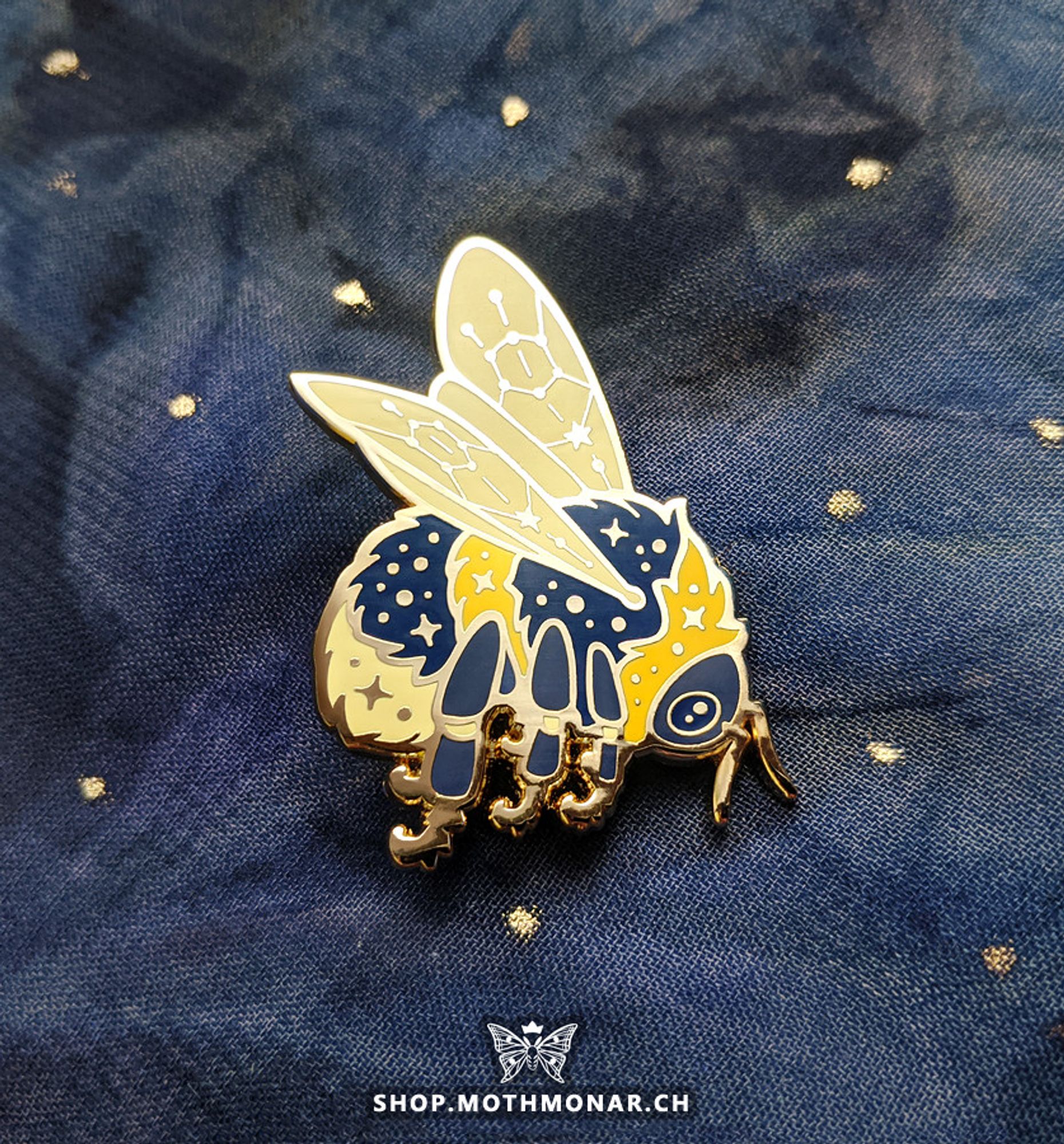 Photo of an enamel pin of a celestial-themed bumblebee. The bee is facing right in a rounded pose with wings sticking out above it. It has a large round eye and sparkles on the body with constellation-like patterns as the wing veins. The antennae and some of the legs are mostly depicted by gold metal. The pin is resting on a blue cloth surface with some round gold accents. Mark at the bottom of the image reads "shop.mothmonar.ch"