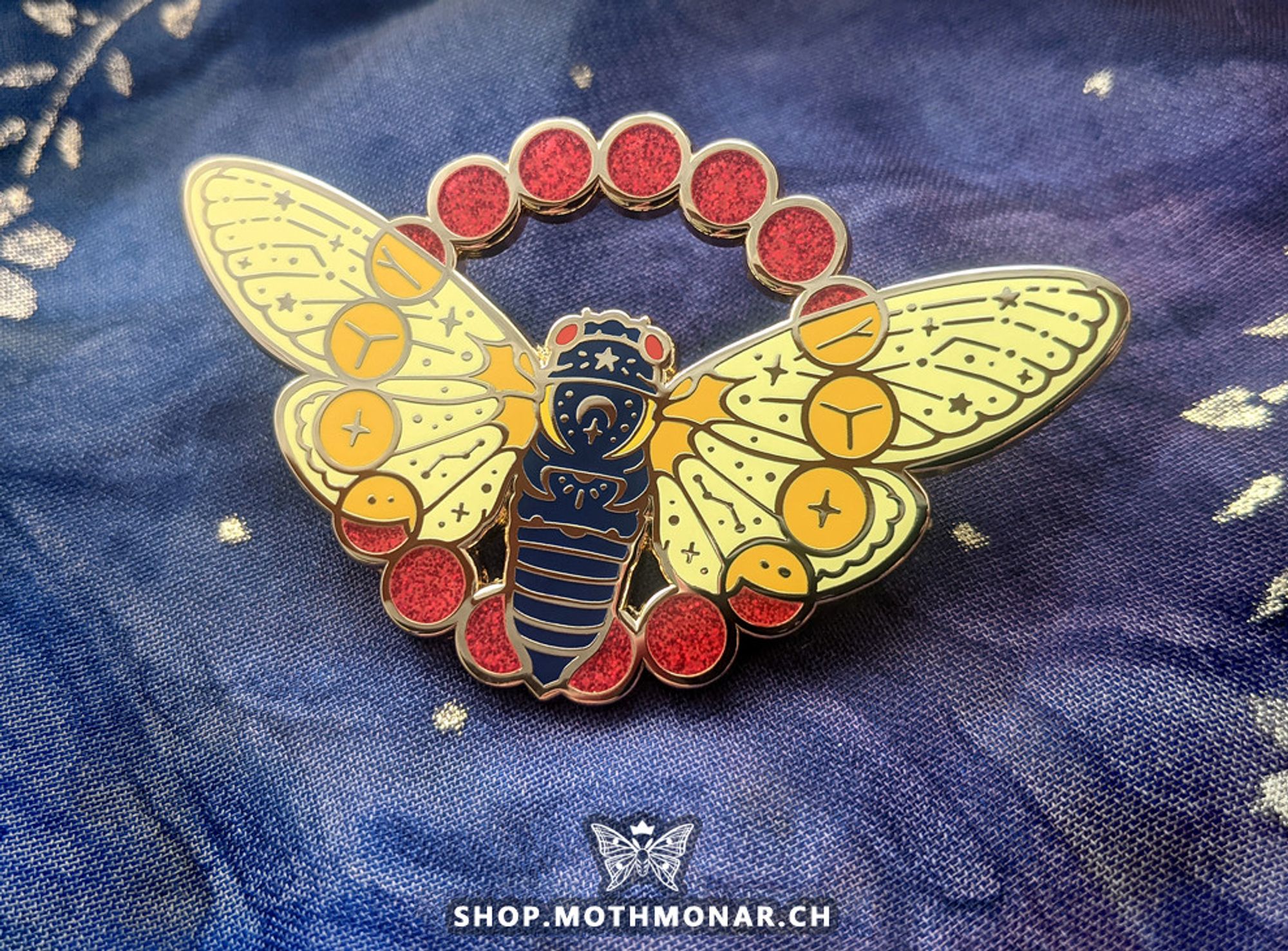 Photo of an enamel pin of a celestial-themed cicada. The insect is seen from above with a ring of 17 circles for suns incorporated over the spread wings. The body is a dark blue, with pale yellow wings and red glitter in the suns where they do not overlap with the wings. It has a few stars, a crescent moon, and some small rays incorporated into its upper back as well as constellation-like veins on its wings. The pin is resting on a blue and purple cloth surface with some gold accents. Mark at the bottom of the image reads "shop.mothmonar.ch"