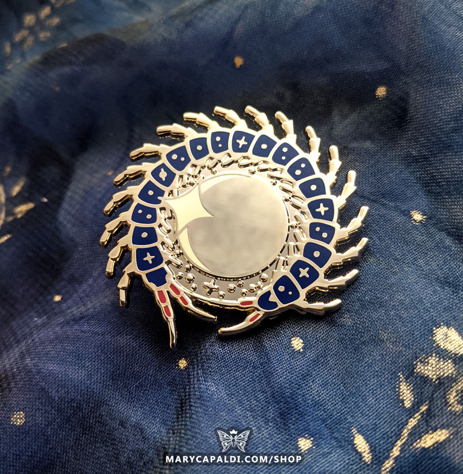 Photo of an enamel pin design of a centipede with celestial markings on its body segments wrapped around a stylized eclipse. The backdrop of the pin is blue fabric with gold details.