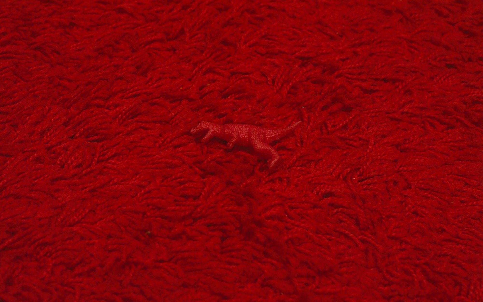 Almost perfectly camoflaged small red squishy rubber dinosaur toy on a red rug.