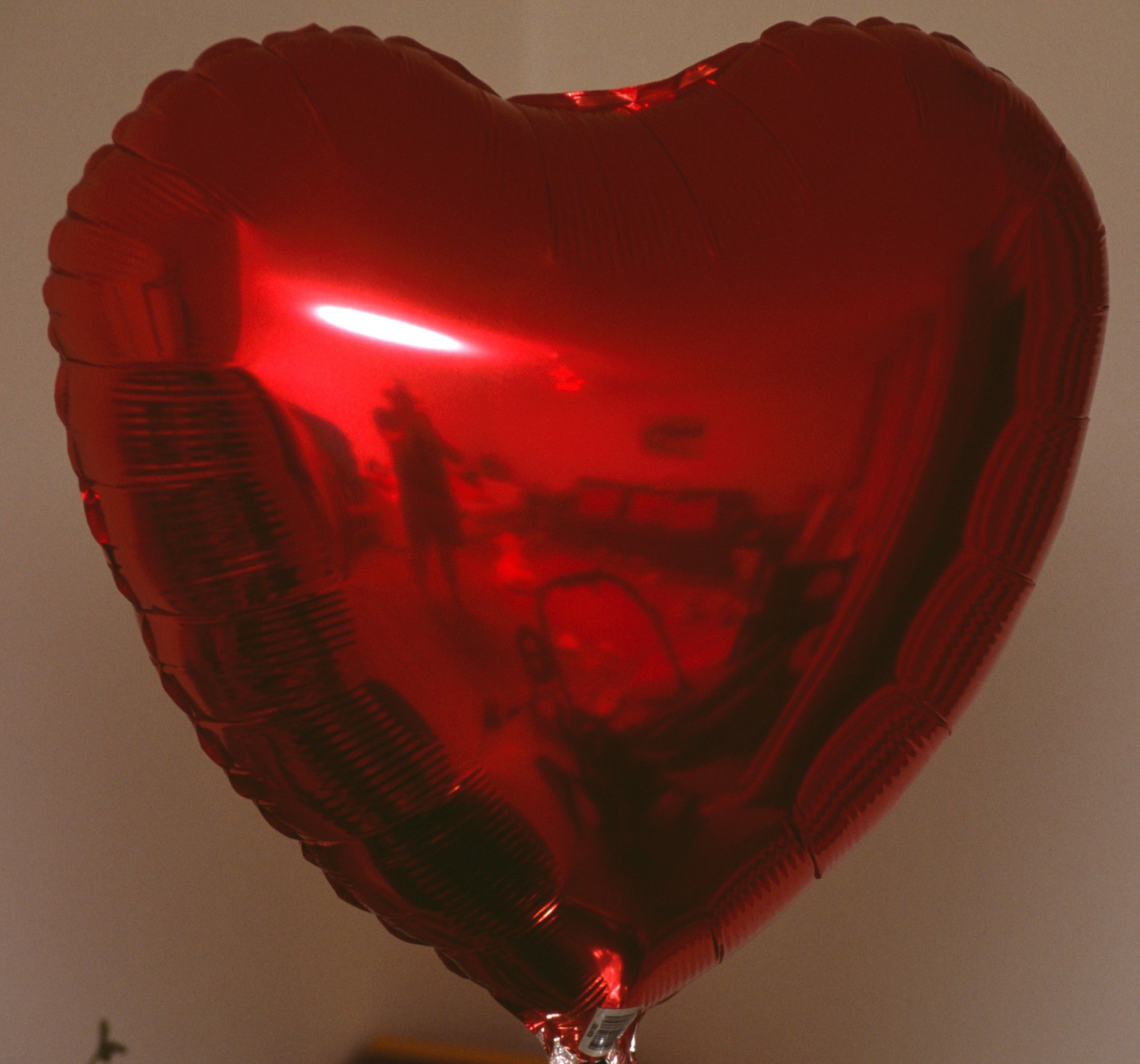 Red, heart shaped helium balloon. The surface of the balloon acts as a mirror showing a distorted view of the photographer and the childrens toys scattered accross the floor of the room.