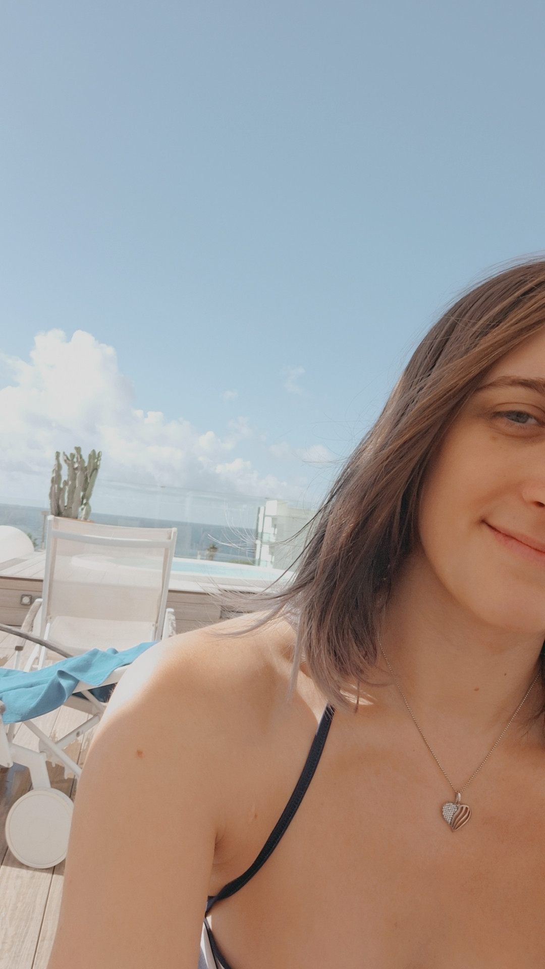 Top of the hotel, showing the blue sky, a jacuzzi and a little bit of ocean in the background. 
Kaylia is taking a selfie, showing half her front with a soft smile. 