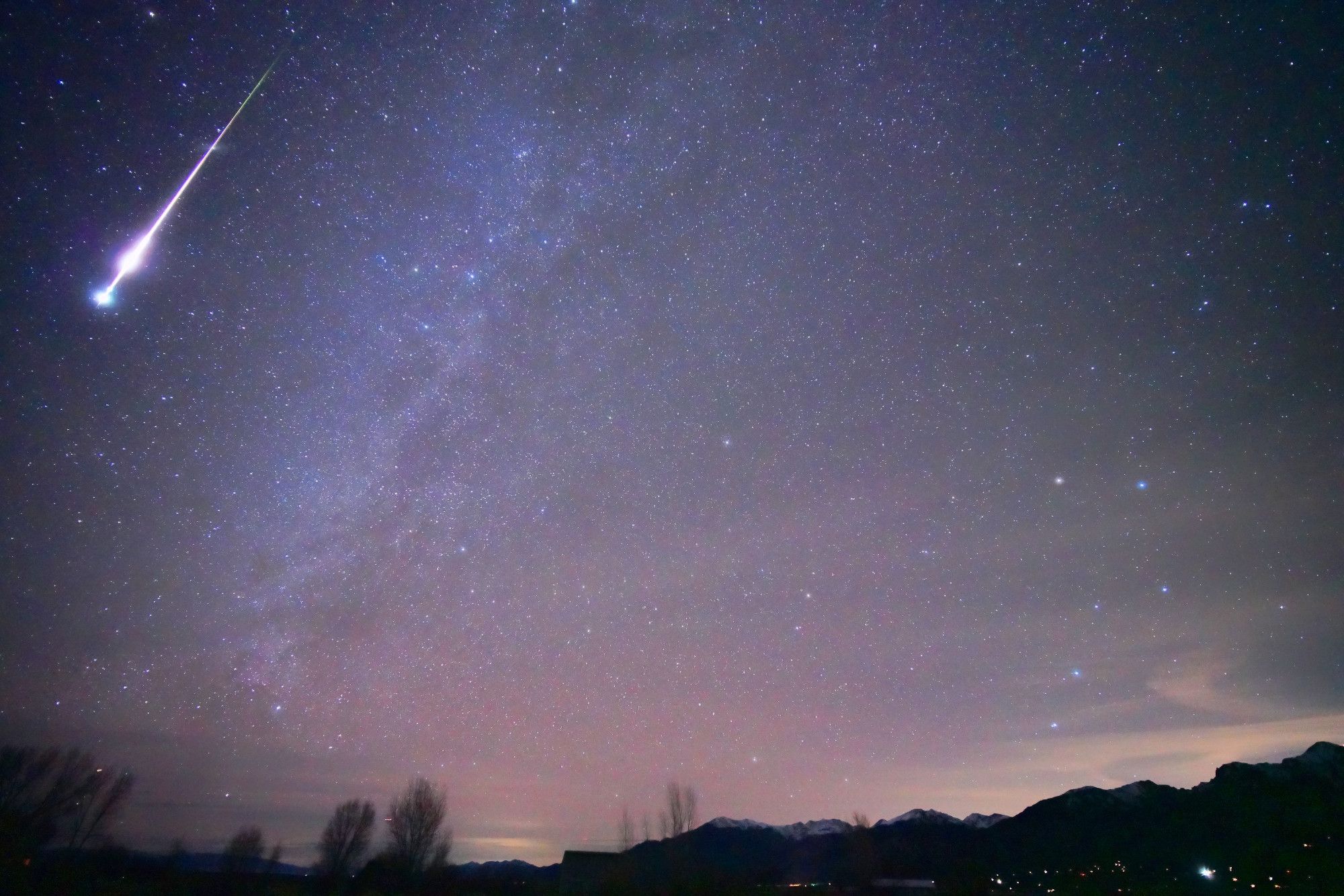 A bright, multicolored meteor streaks down in the upper left corner. The tail is green, turning to purple, then a pink flare, narrowed down again and finally terminating in a slightly blue bulb. The Milky Way is to the left of the meteor. Ursa Major, the Big Dipper, is visible on the right. Polaris and the Little Dipper Ursa Minor are centered.