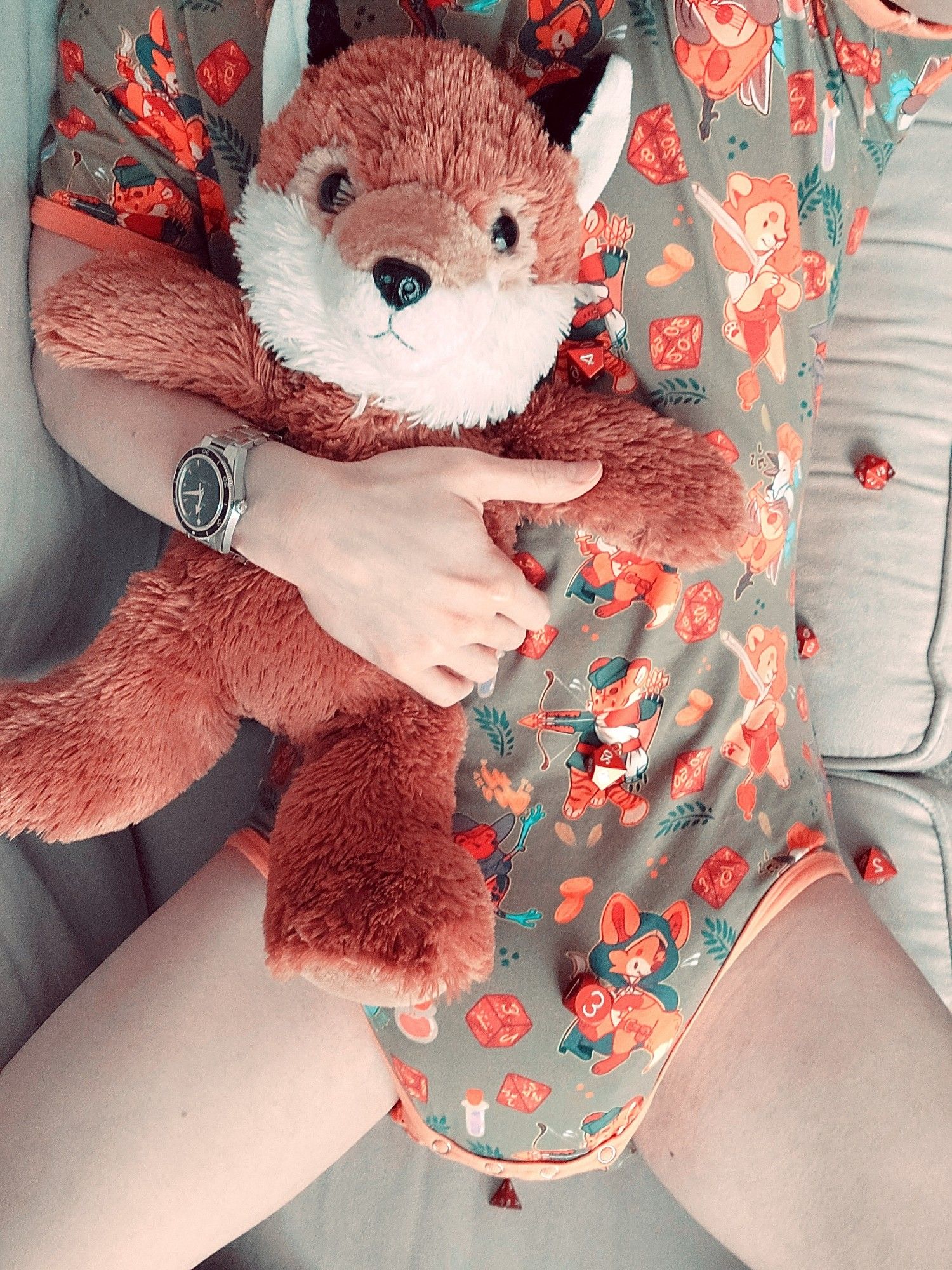 Me laying on the couch, snuggling my plush fox. I'm wearing a green and orange onesie with a D&D themed print. There are dice scattered about.