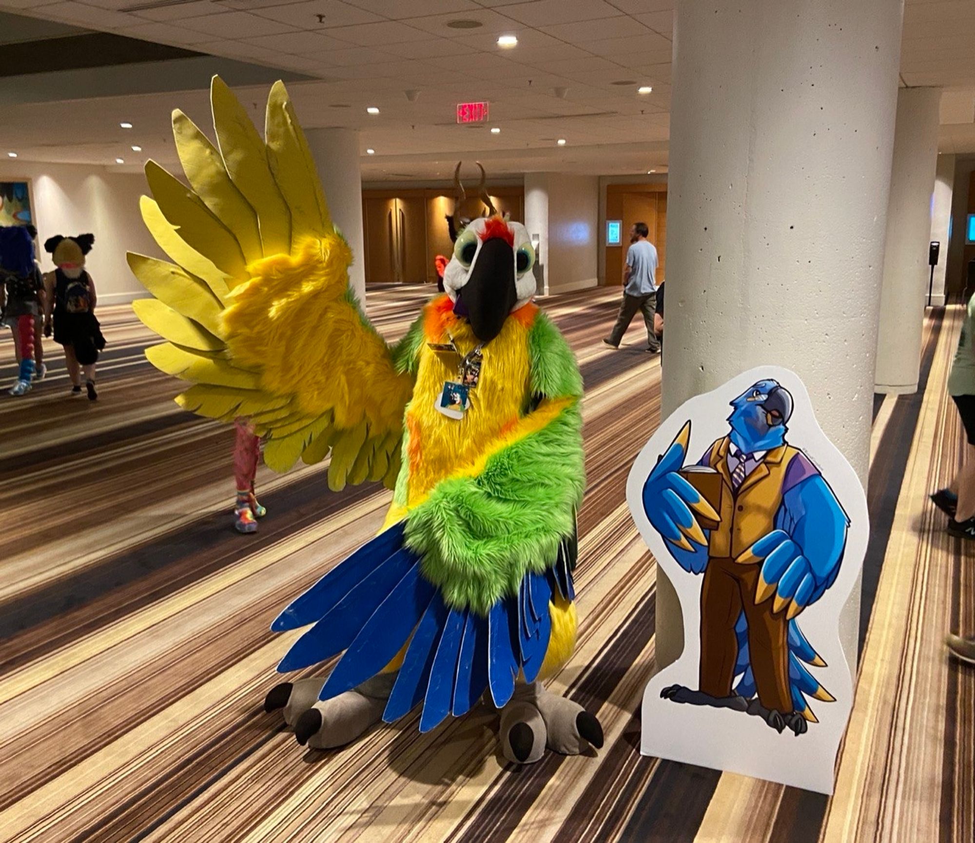 Carl Lee the macaw at FWA 2024 next to a Hyacinth Macaw professor standee. Carl Lee is trying to mimic his pose with one wing raised and the other in front of him.