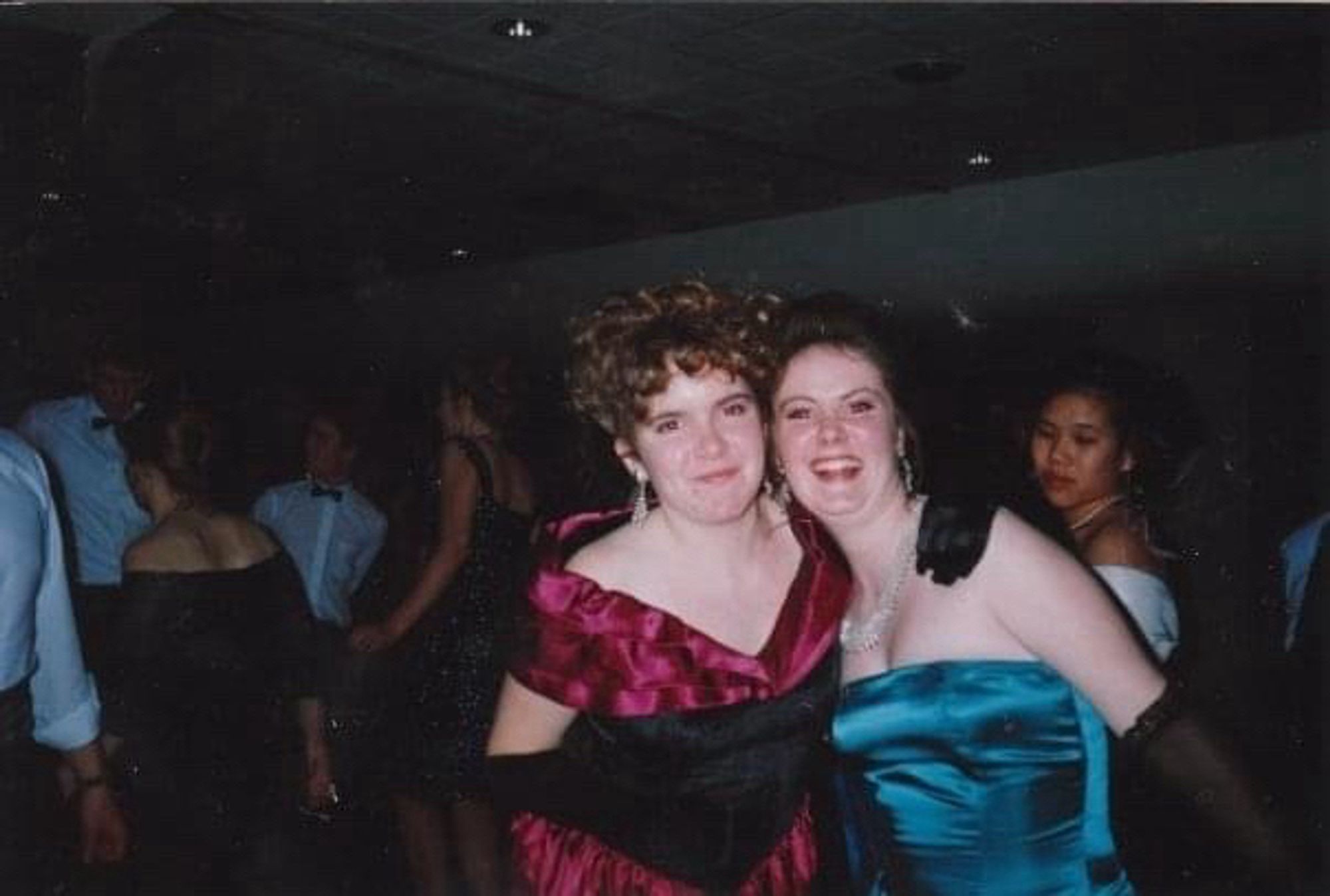 Two young women in ball dresses, black gloved arms around each other’s shoulders and grinning on the dance floor