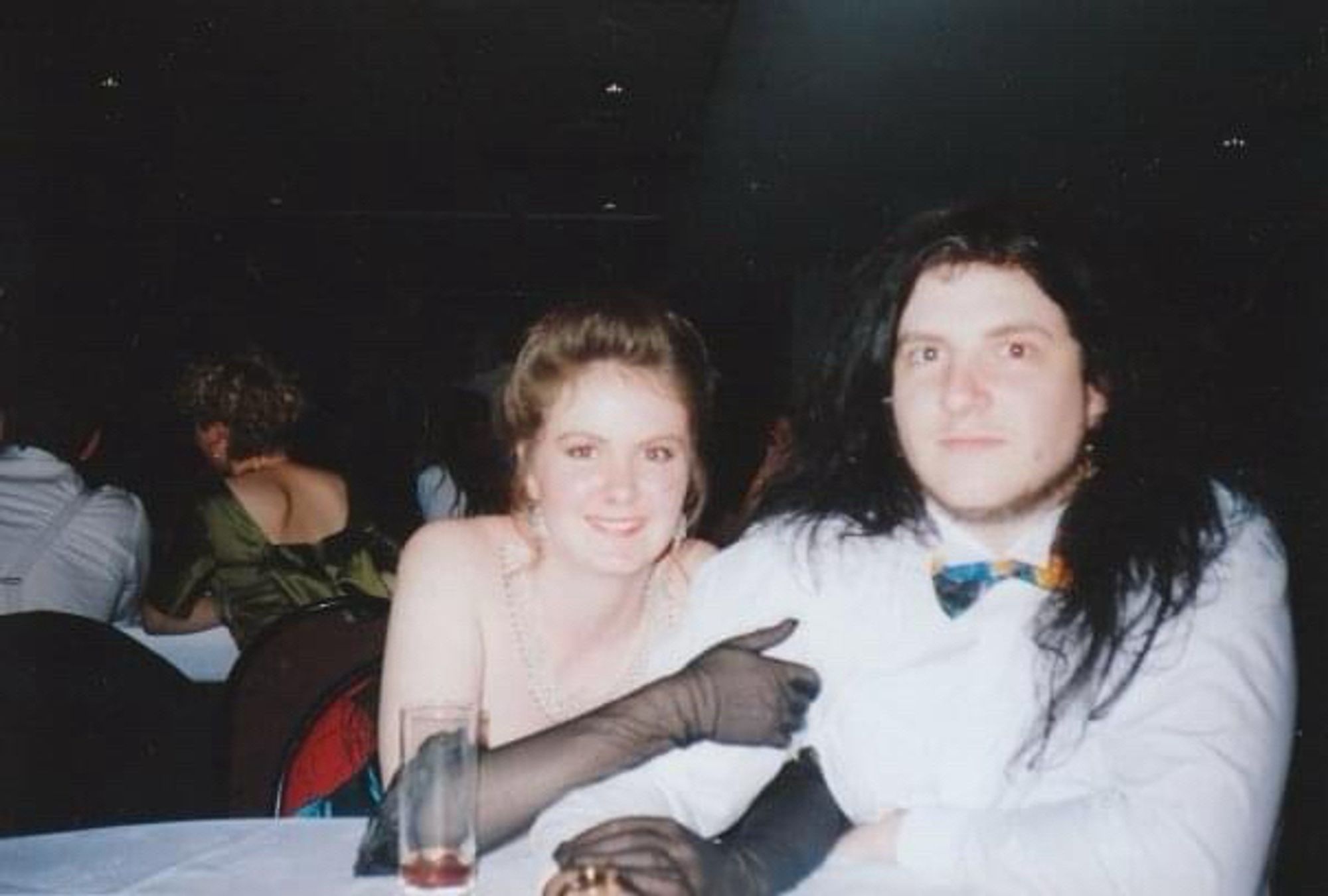 A young woman in a strapless ball gown sits at a table next to a young man with long black hair and a multi-coloured bow tie