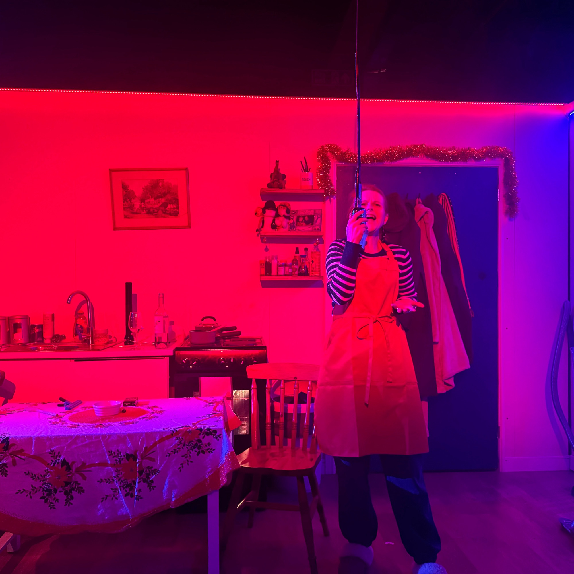 Cathy speaks into a microphone hanging from the ceiling. She is wearing a red and green stripey top with a red blue joggers, and some moccasin slippers. She is standing in a kitchen that is lit with blue and red light.