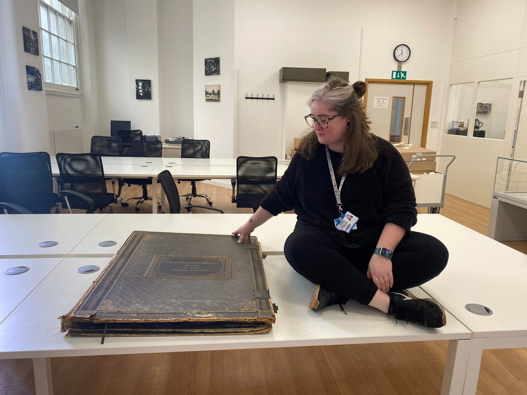 Woman wearing all black sitting cross legged on a table next to a large book of plans