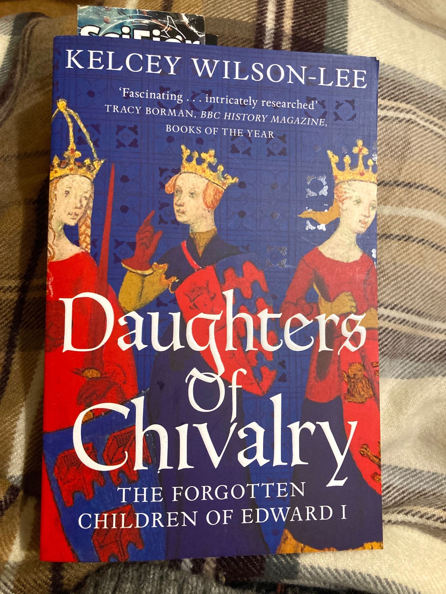 A paperback of Daughters of Chivalry by Kelcey Wilson-Lee. The subheading of the book reads: “The forgotten children of Edward I”. The cover shows a 13th century illustration of three young women wearing golden crowns and carrying shields and swords.