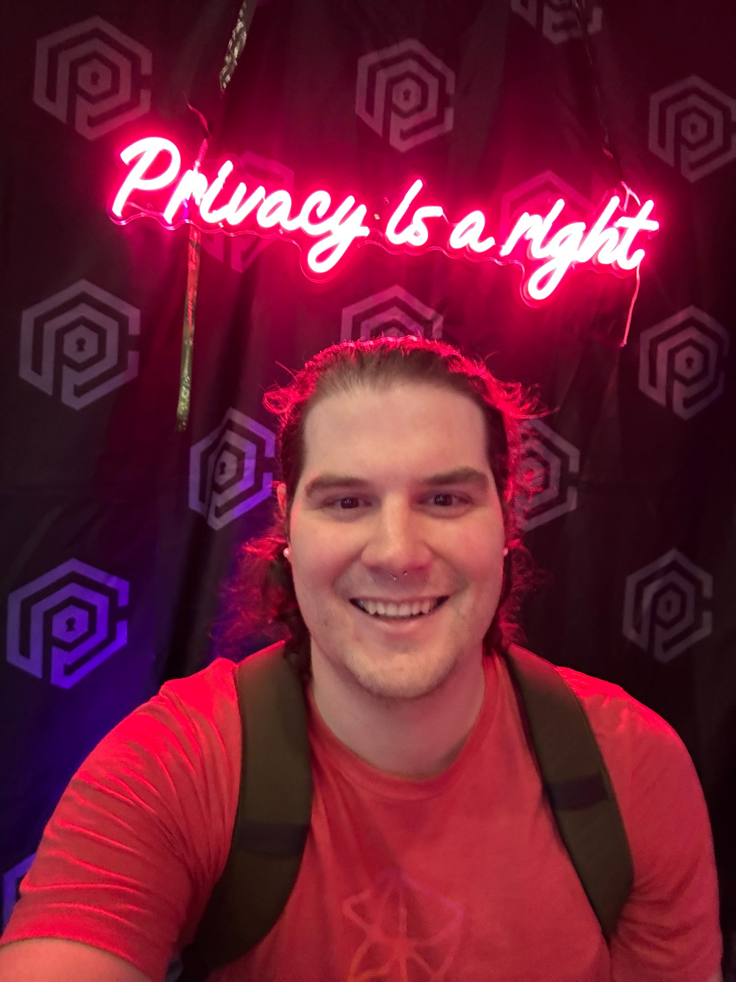 Me - a white masculine person with long hair and a Zune t shirt in front of a neon sign with scripty font that reads “privacy is a right”