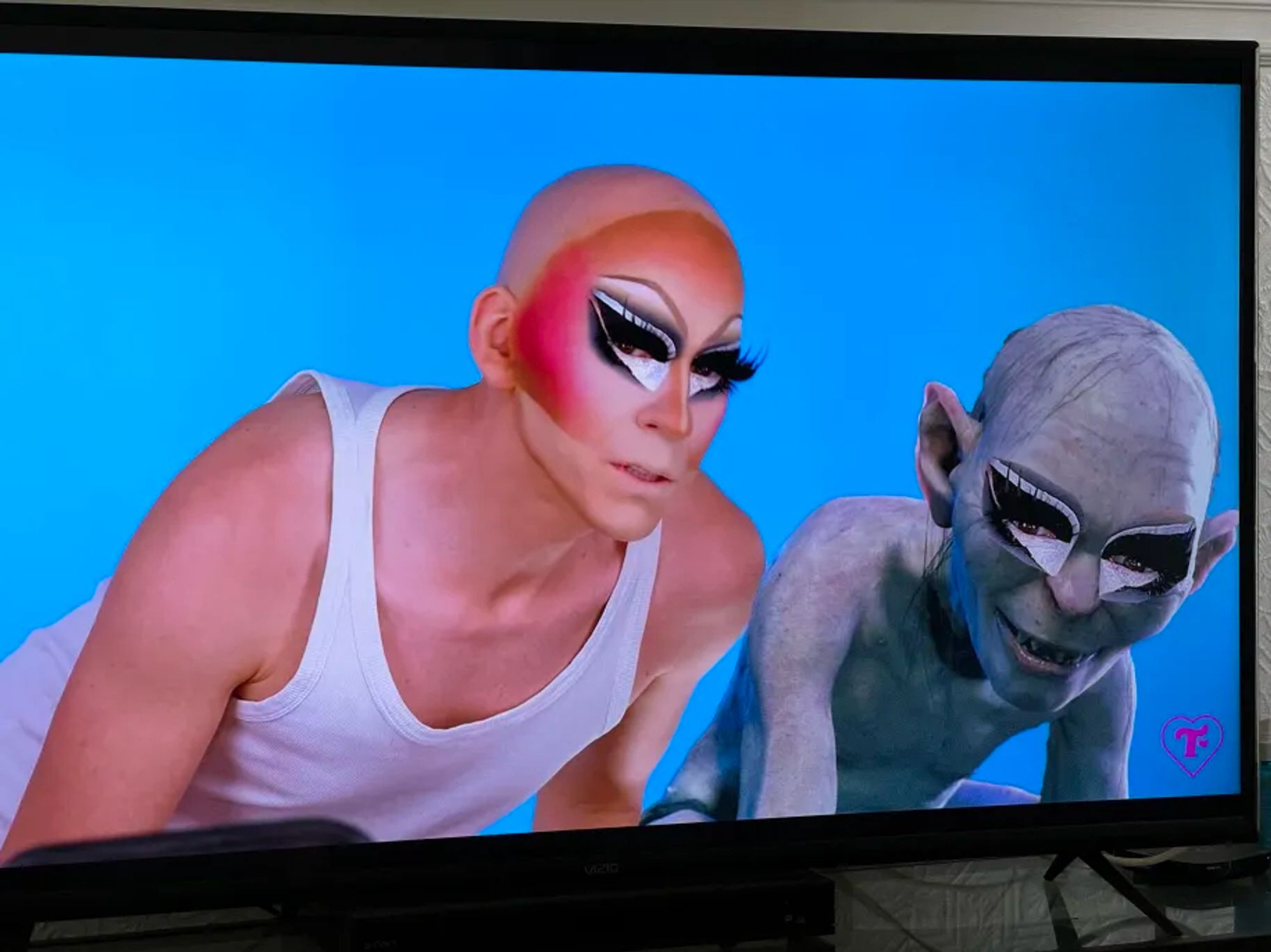 A shot from Trixie Mattel's latest YouTube channel entry showing her with her face partially on but no wig wearing a white tank top, standing slightly up at an angle. Superimposed on the right is a pose of Gollum from the LOTR films at a similar angle, with Trixie makeup on.