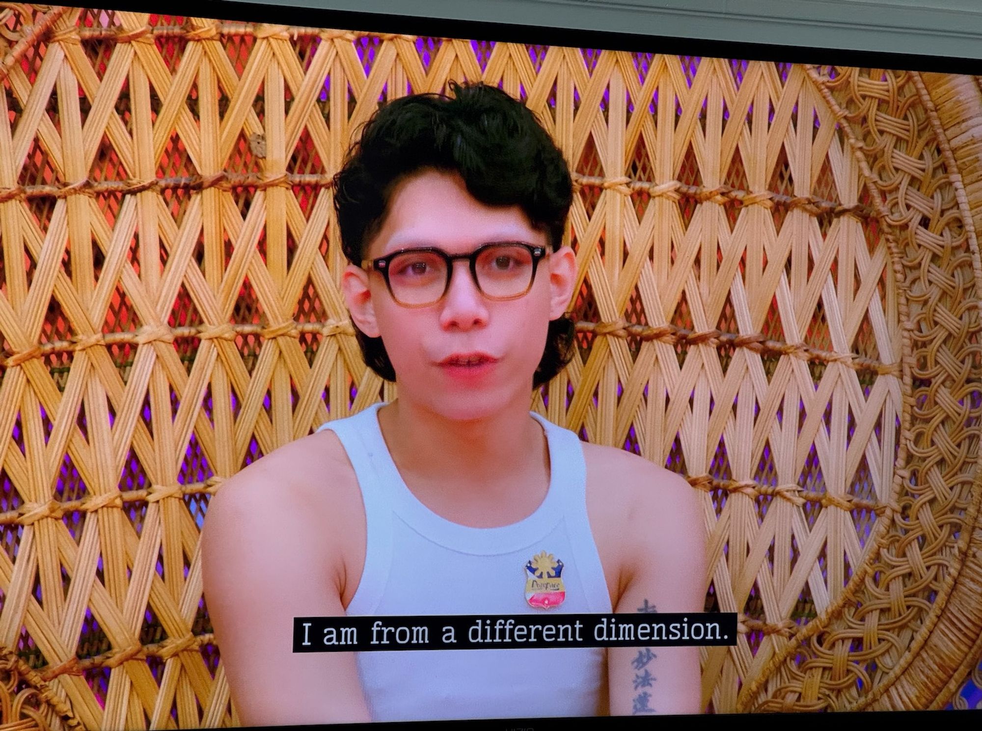 In the Drag Race Philippines werk room, Khianna with a K, sitting in a rattan chair in boy look with glasses and a white tank top, says to the offscreen Drag Council "I am from a different dimension."