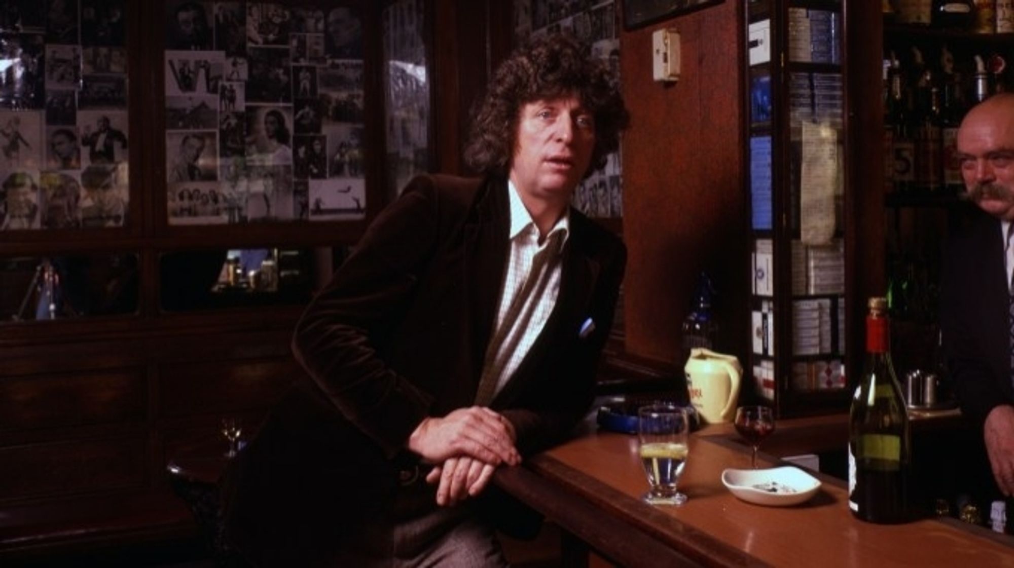 A clearer version of the picture used in the printed article from the original post. Tom Baker is leaning against the barbof an old London pub, looking contemplative, nursing a drink, possibly mid-sentence saying something scandalous, with a toothbrush cheekily sticking out of his top pocket. Behind him are several black and white photos on the wall, likely of famous clientele of the day.