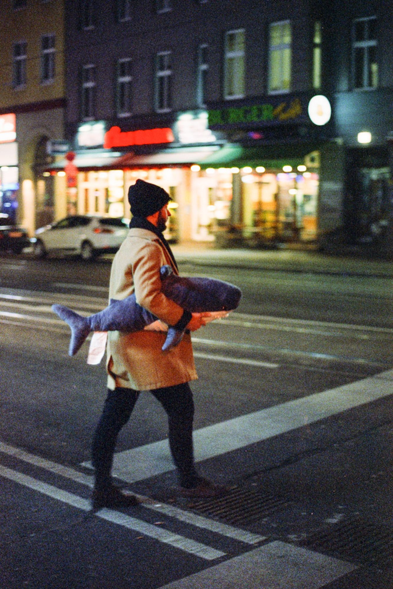 Das Bild zeigt eine Person, die bei Nacht über eine Straße geht. Sie trägt einen beigen Mantel und eine Mütze, während sie in den Armen einen großen Plüschhai hält. Der Hai ist detailreich und auffällig, was dem Bild eine humorvolle Note verleiht. Die Person geht in einer urbanen Umgebung, möglicherweise in einer geschäftigen Straße, die von den Lichtern der Geschäfte und Restaurants im Hintergrund beleuchtet wird. Die nächtliche Atmosphäre und die unscharfen, bunten Lichter erzeugen ein warmes, lebhaftes Stadtbild, das durch den ungewöhnlichen Begleiter – den Plüschhai – kontrastiert wird.