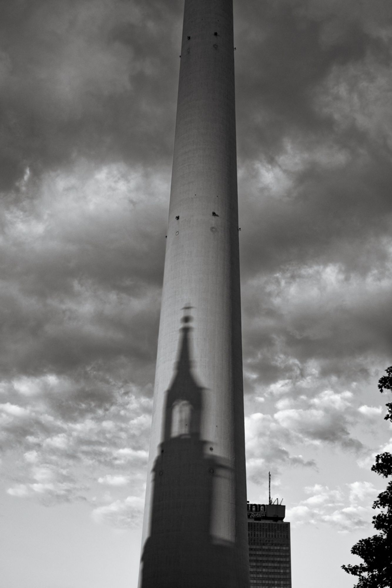 Das Bild zeigt den unteren Teil eines hohen Turms, auf den der Schatten eines Kirchturms projiziert wird. Der Turm ist schlicht und scheint aus Metall zu bestehen, während der Schatten einen traditionellen Kirchturm mit spitzer Spitze und einem rechteckigen Glockenturm zeigt. Im Hintergrund sind dramatische Wolken am Himmel zu sehen, die dem Bild eine düstere und geheimnisvolle Atmosphäre verleihen. Rechts im Bild ist ein Baum oder ein Strauch zu erkennen, und im Hintergrund steht ein hohes Gebäude mit einer Antenne auf dem Dach. Die Komposition des Bildes betont den Kontrast zwischen moderner Architektur und traditioneller kirchlicher Architektur.