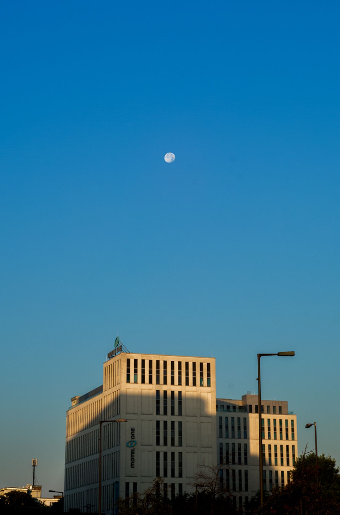 Das Bild zeigt einen klaren blauen Himmel mit dem Mond, der hoch über einem modernen, rechteckigen Gebäude steht. Das Gebäude ist ein "Motel One", das im unteren Bereich des Bildes platziert ist und von der tief stehenden Sonne in goldenes Licht getaucht wird. Die warme Beleuchtung auf dem Gebäude kontrastiert stark mit dem kühlen Blau des Himmels. Einzelne Straßenlaternen sind ebenfalls zu sehen, wobei das Bild eine ruhige, minimalistische Atmosphäre erzeugt, die den Mond und das Spiel aus Licht und Schatten in den Fokus rückt.