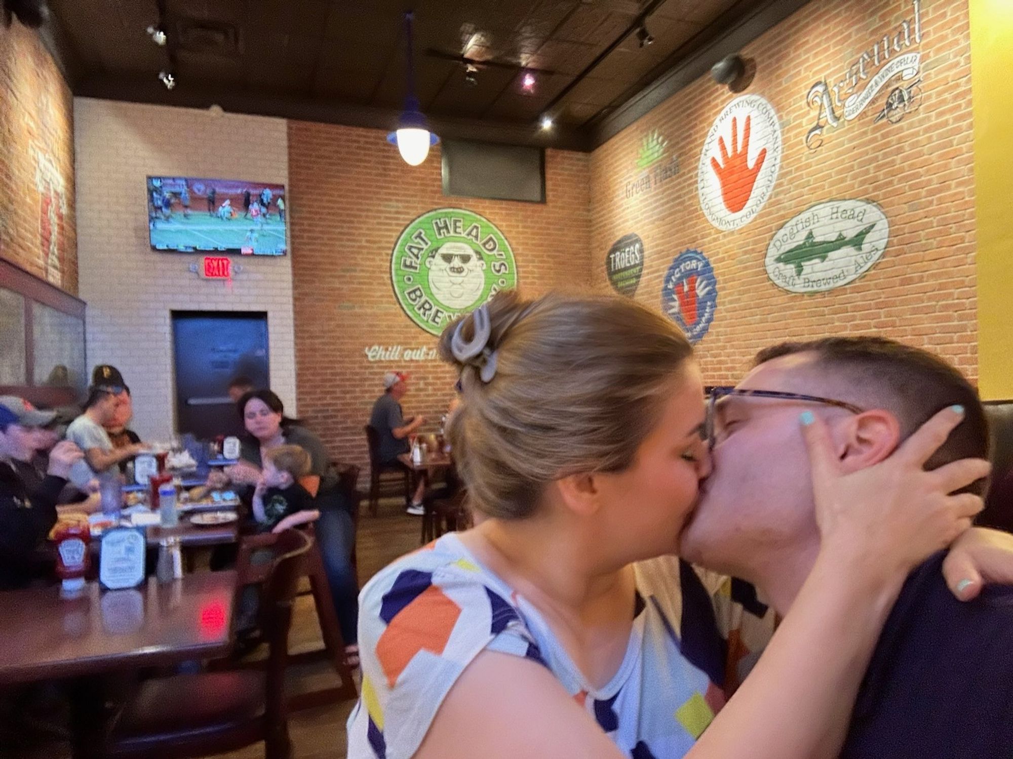 Me and my husband kissing each other in a bar with a brick wall that notably has the Fatheads logo behind our macking heads