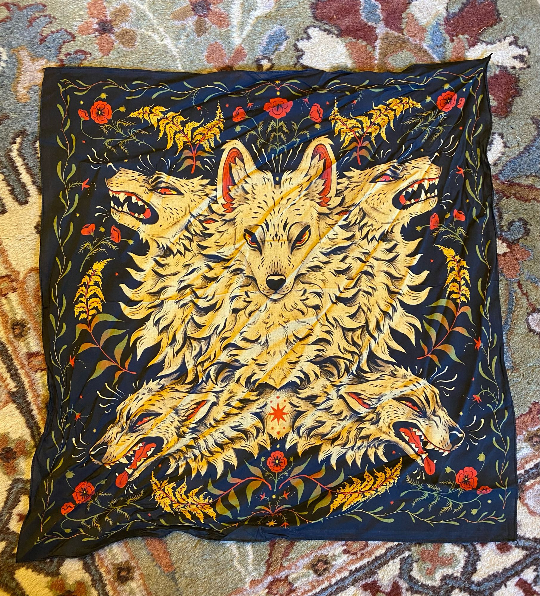 A bandana laid out on the floor, featuring art by Mothsprout. It is black with five off-white canine heads fanned out in the center, with yellow and red flowers and green leaves circling them.