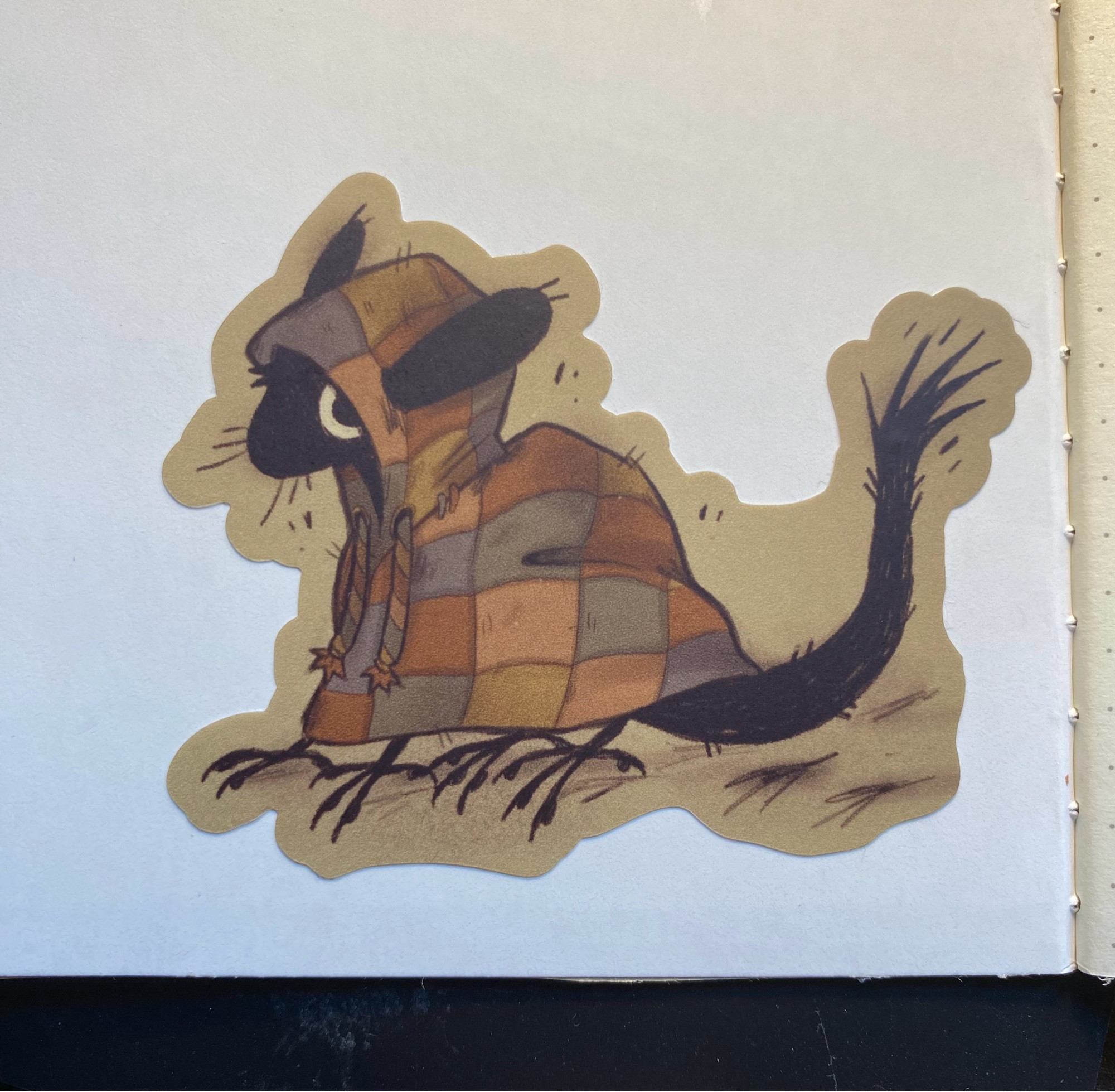 A small sticker stuck on the corner of a journal of a black soot creature. It has birdlike feet, a mouselike head, and a long tufted tail. It's wearing a multicolored hoodie made from squared of fabric