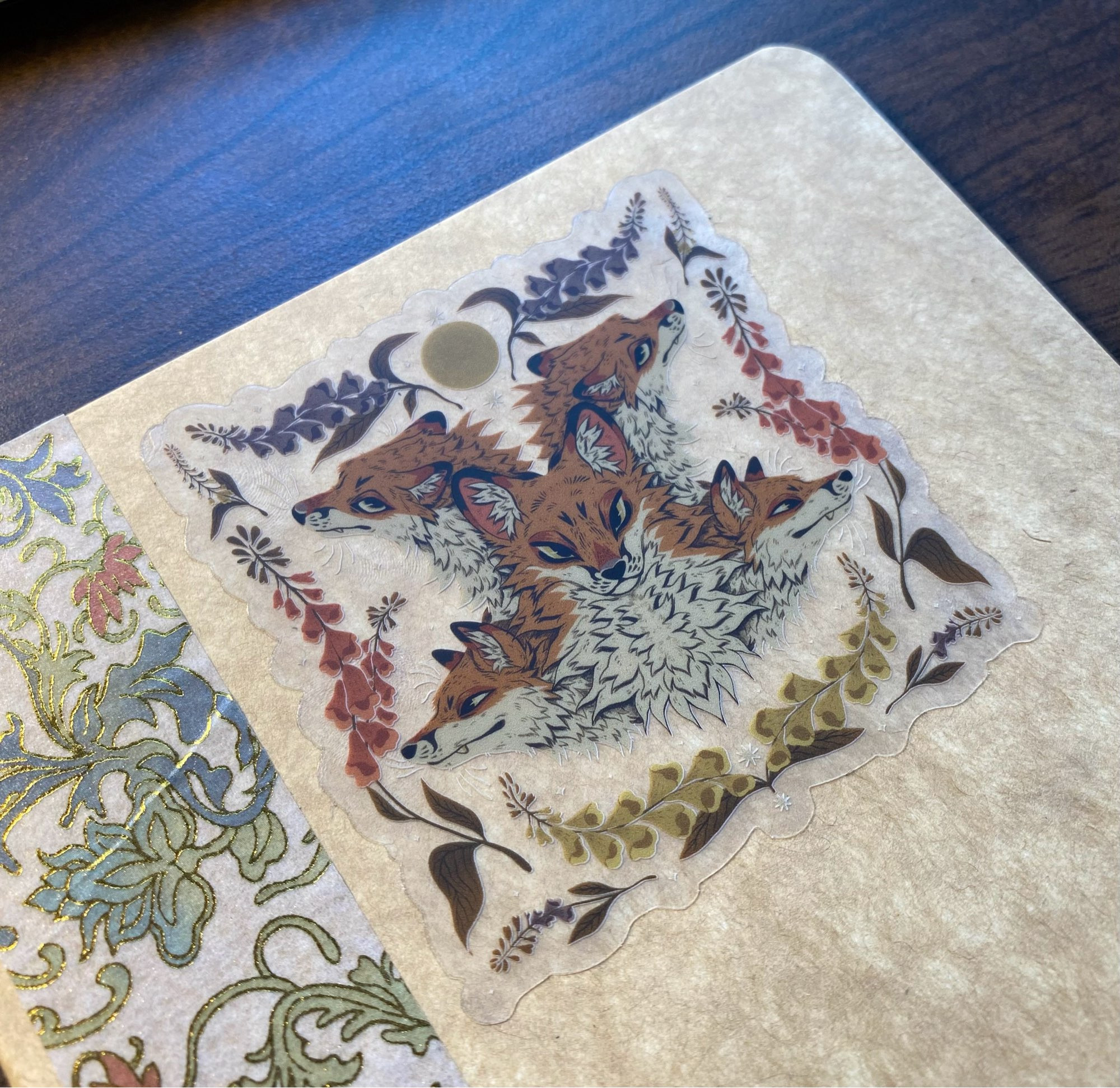 A sticker with four fox heads fanned out in a circle and a fifth one in the center. Purple, yellow, and pink flowers surround them. The back of the sticker is clear and you can see the texture of a tan journal cover through it