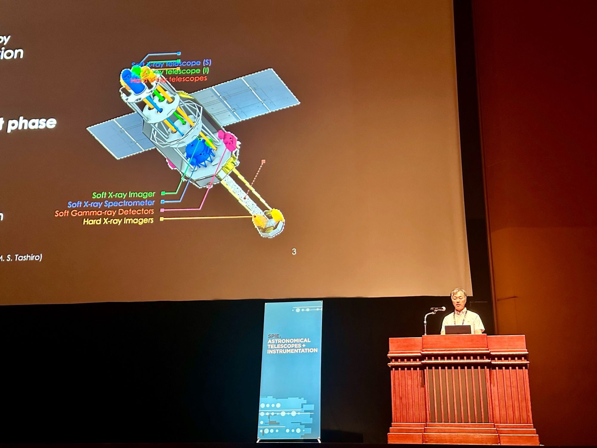 A speaker is presenting on stage in an auditorium. The slide behind the speaker shows a detailed diagram of a space telescope with various labeled components, such as "Soft X-ray Imager," "Hard X-ray Imagers," and "Soft Gamma-ray Detectors."