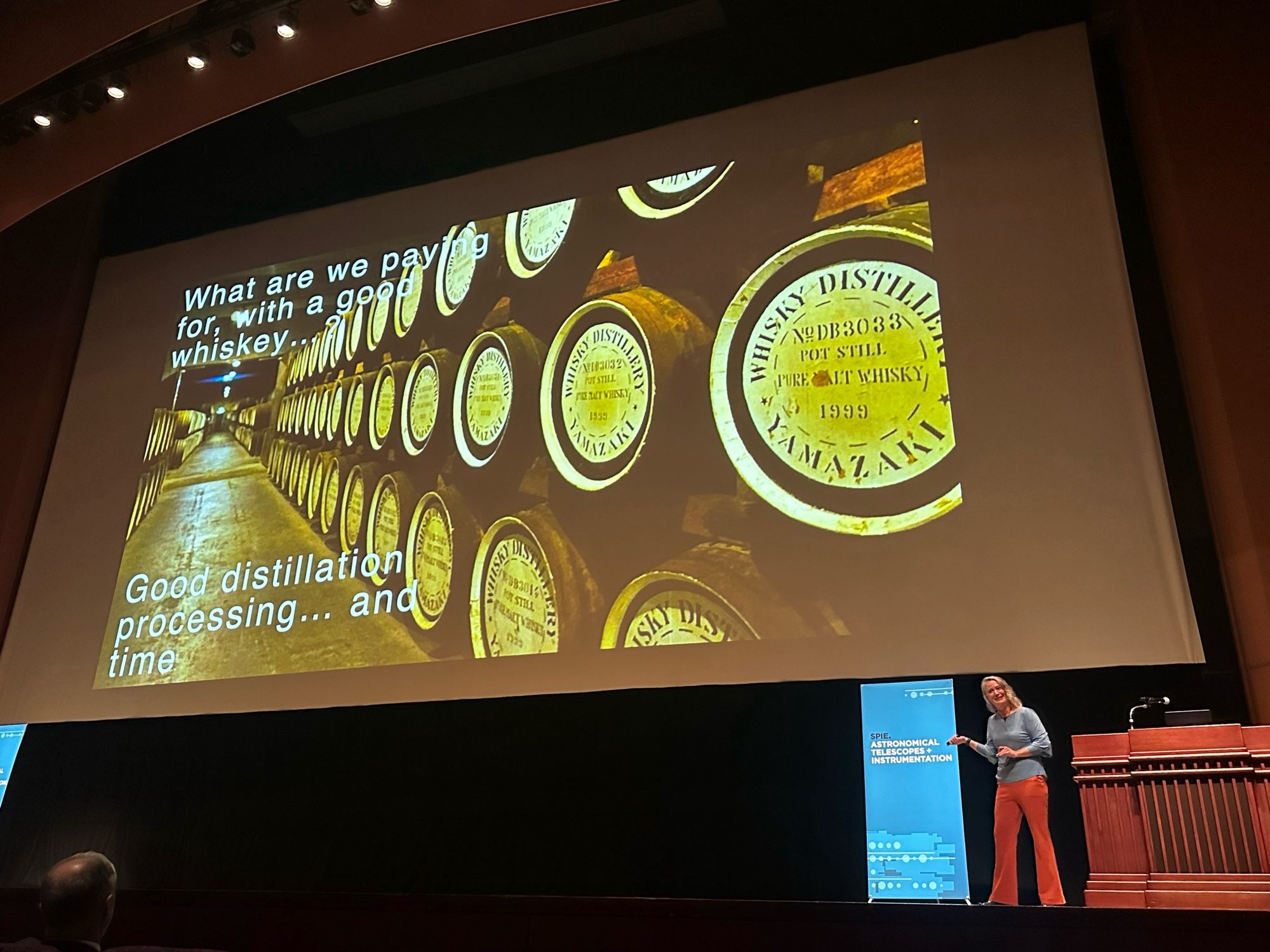 A speaker wearing a blue shirt and orange pants is presenting on stage in an auditorium. The slide behind the speaker shows a photo of whiskey barrels with the text "What are we paying for, with a good whiskey?" and "Good distillation processing... and time."