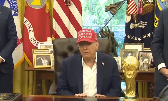 Donald Trump sitting at the oval office desk surrounded by pictures, flags, statues, trophies, etc. 