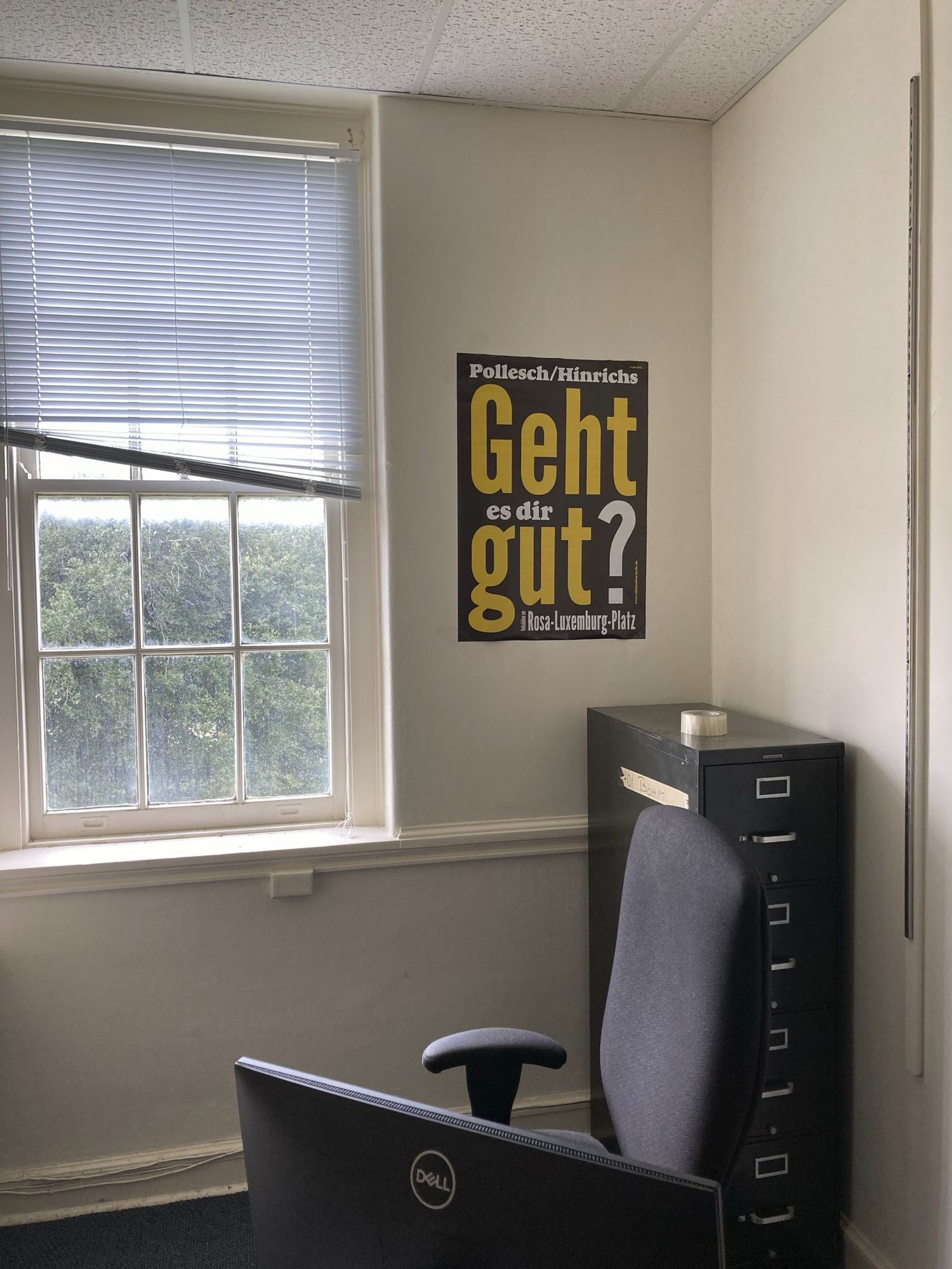 Heruntergekommene Büroecke: Fenster mit kaputter Jalousie, Aktenschrank und ein schwarz-gelbes Poster der Theaterinszenierung "Geht es dir gut?" (Volksbühne Berlin, René Pollesch/Fabian Hinrichs)