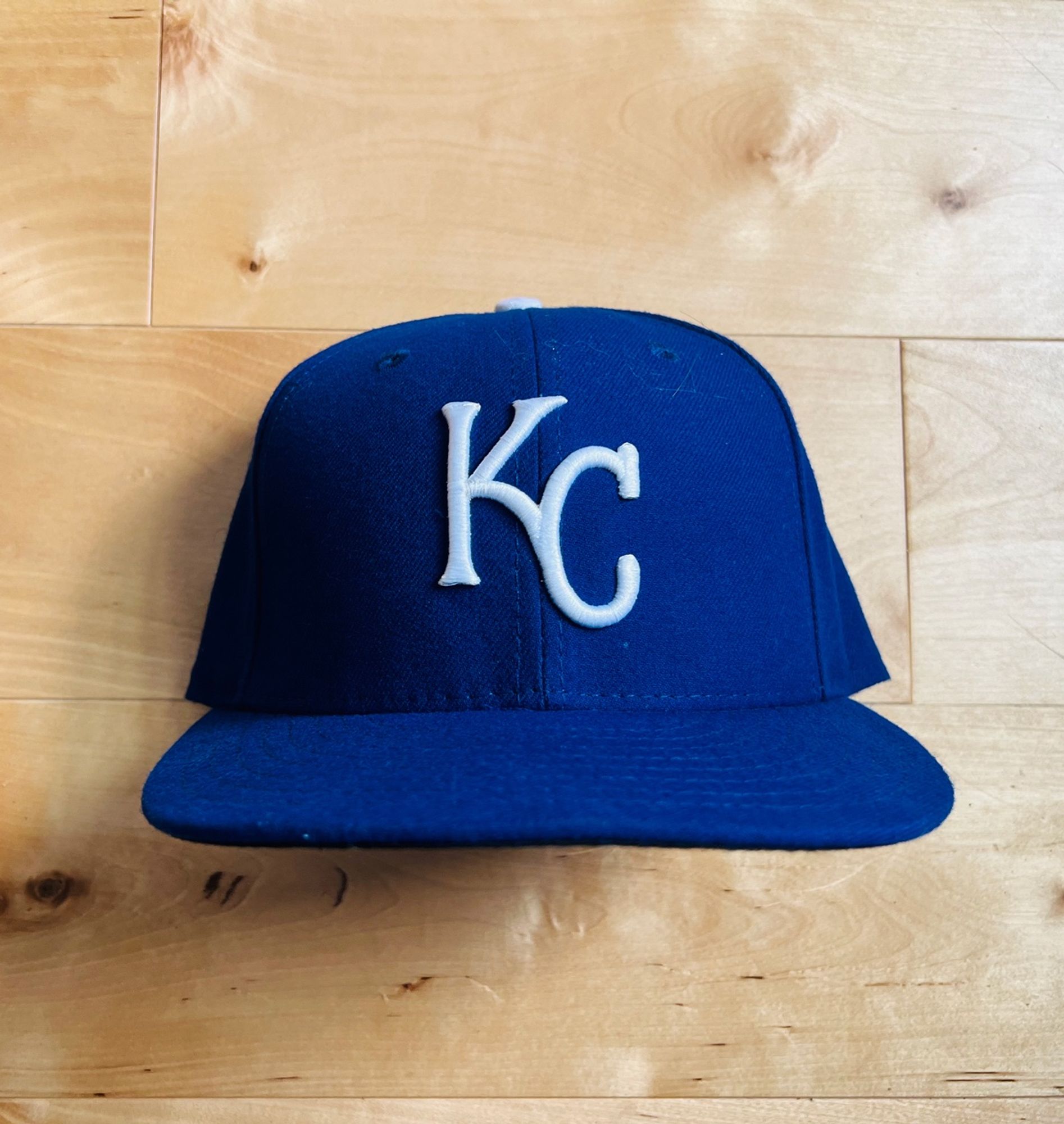 The Kansas City Royals home hat. They've worn it the entirety of their time as a club. The hat features a blue crown and visor. There's a white button. The logo is a "KC" monogram in which the C extends from the lower right of the K.