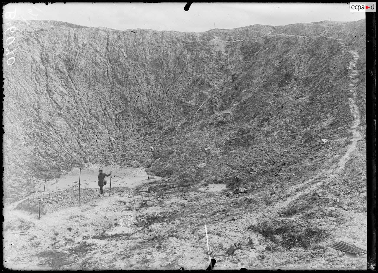 Black and white photo of the interior of an enormous mine crater, at the bottom of which a man looks at a rectangular mound on which can be seen a skull, bones and shell casings. 
© Paul Queste/ECPAD/Défense SPA 114 B 6070
https://imagesdefense.gouv.fr/fr/la-boisselle-somme-entonnoir-de-mine-anglaise-28-m-de-profondeur-diametre-60-legende-d-origine.html