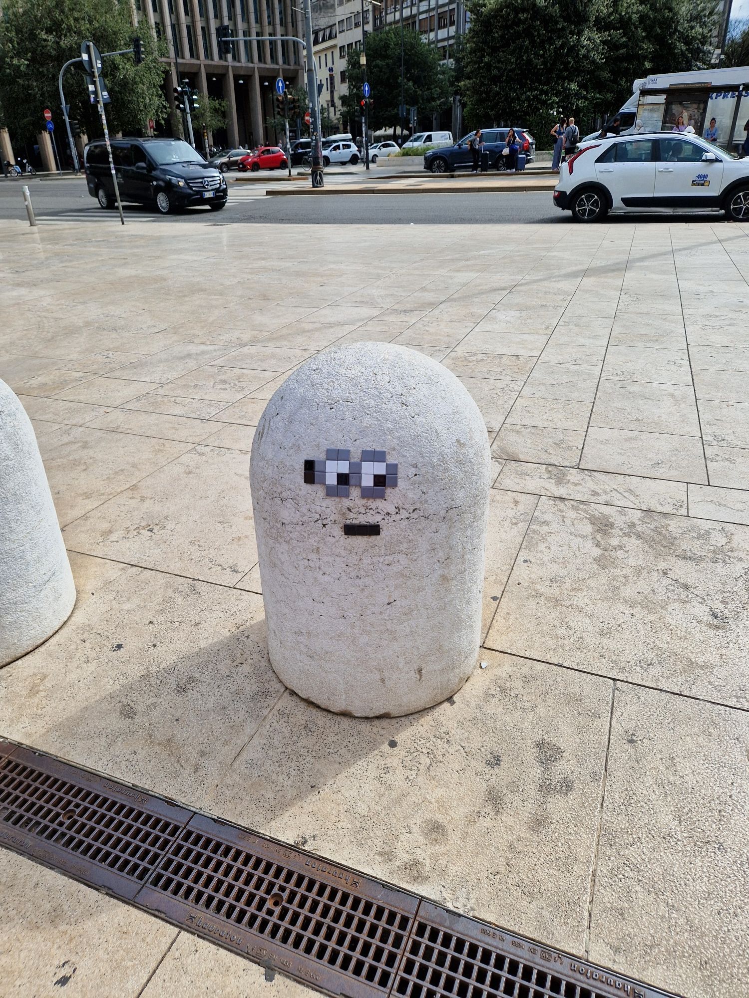 Ein kleiner, grauer, oben runder Steinpoller zwischen Gehweg und Weg zum Bahnhof. Er steht auf hellen Gehwegplatten. Jemand hat aus kleinen, quadratischen Patches Augen, die vom Betrachter aus nach rechts unten schauen, und einen Mund raufgeklebt. Die Augen bestehen aus einer grauen Umrandung, weißer Lederhaut und 1 schwarzen Patch als Pupille. Der Mund ist aus 3 als Strich geklebten schwarzen Patches. Im Hintergrund eine Strasse mit Autos, Ampeln, Menschen, Bäume und Gebäude