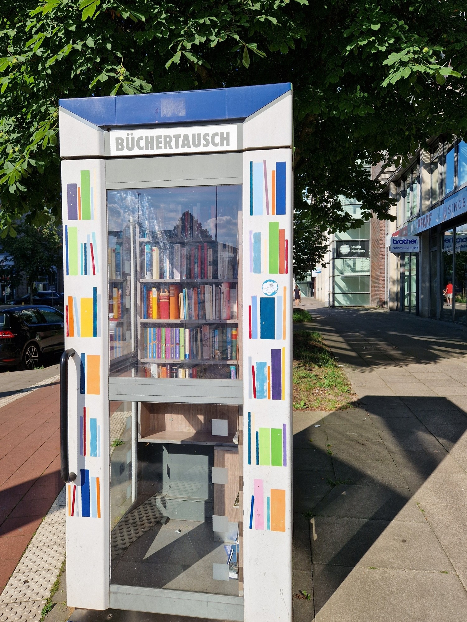 Eine alte Telefonzellenbox mit Tür umgebaut und neu angemalt/beklebt.
Außen auf der Box steht Bücherrausch, links und rechts der Fenster sind bunte Bücher, wie sie in Bücherregalen stehen, aufgeklebt.
In der Telefonzelle sind 6 Regale, 5 davon voll mit Büchern, eins noch leer.
Die Telefonzelle steht auf einem breiten Gehweg rechts neben einem Fahrradweg. Sie steht im Sonnenlicht und wirft einen Schatten nach rechts auf den Gehweg