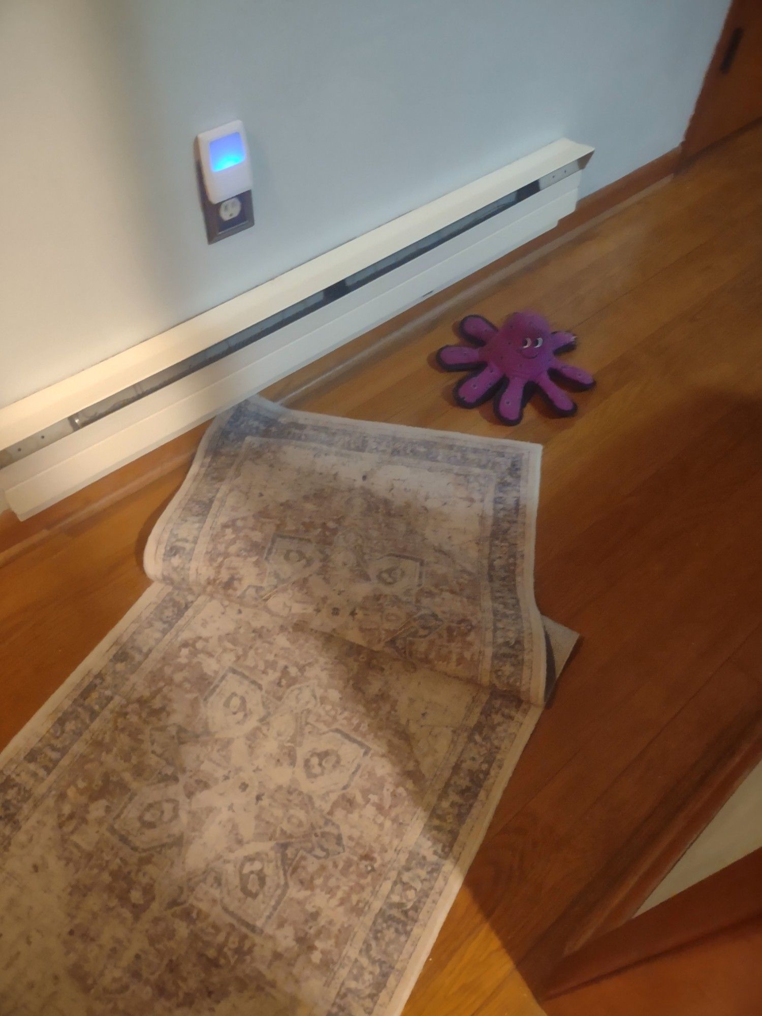 A purple octopus chew toy smiling suspiciously at a disheveled throw rug on a hardwood floor.