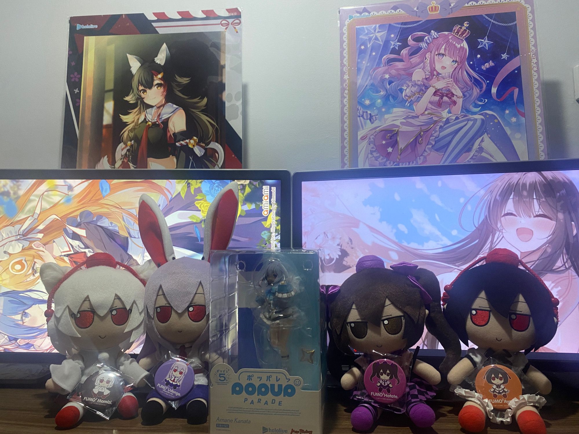 A photo of 4 Fumos and a figure sitting in a line next to eachother. In order, Momiji, Reisen, Amane Kanata Figure, Hatate then Aya. Each Fumo is holding their respective pin. They are sitting in front of two computer monitors, the left depicting the most recent AmiAmi postcard Illustration. While the left shows an illustration by piyopoyo.