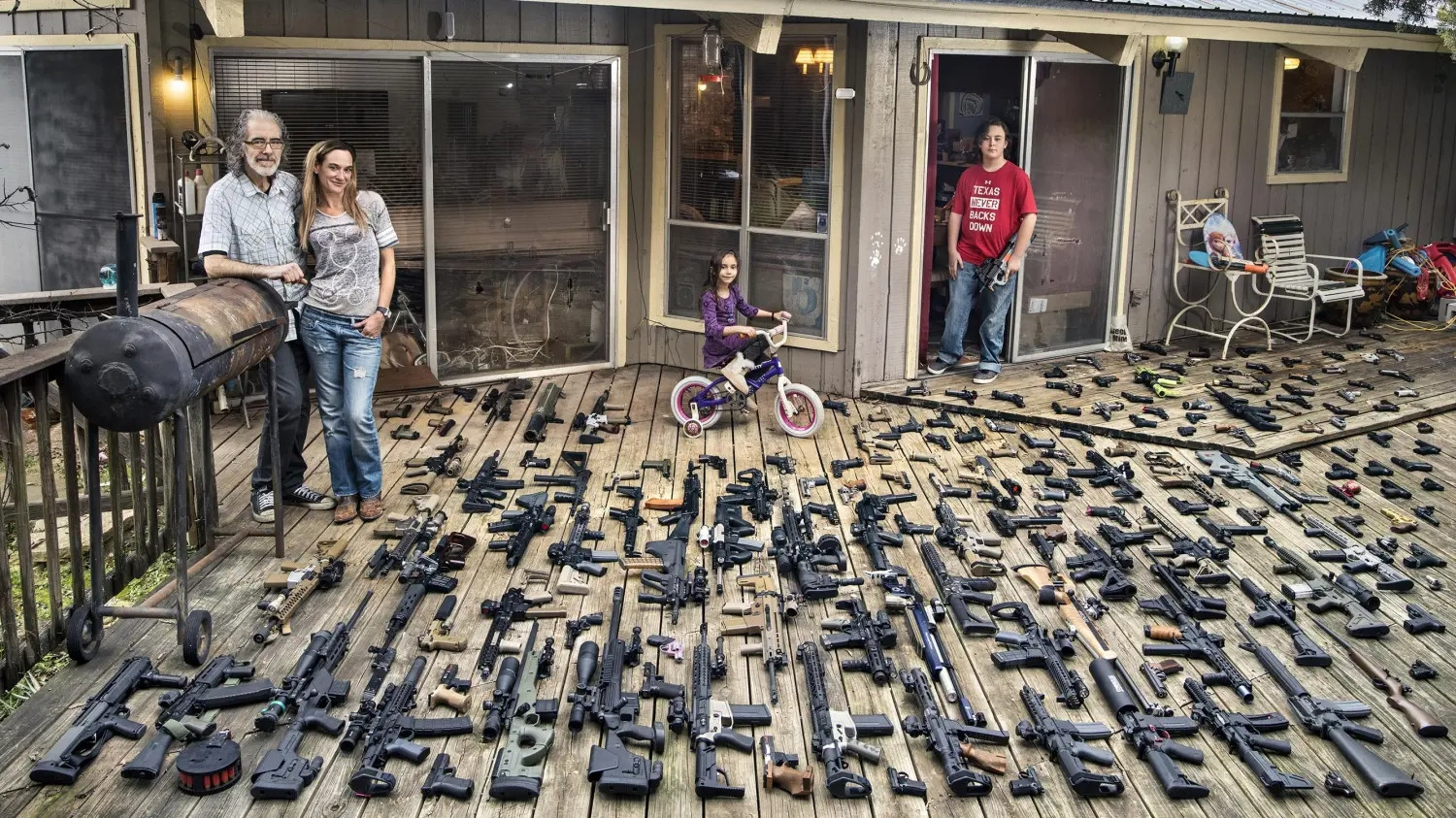A family outside their home. Their deck is loaded with guns. 