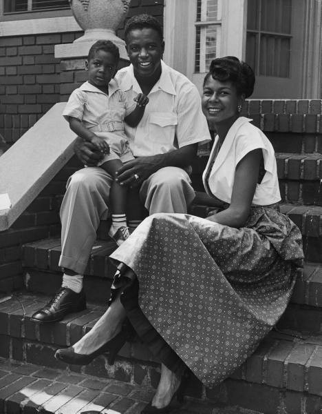 A mid-century family sitting on the porch steps. The man is wearing a camp collar shirt with a flapped pocket (collar and flap have a distinctive shape). he pairs them with fuller cut pants and leather shoes. 