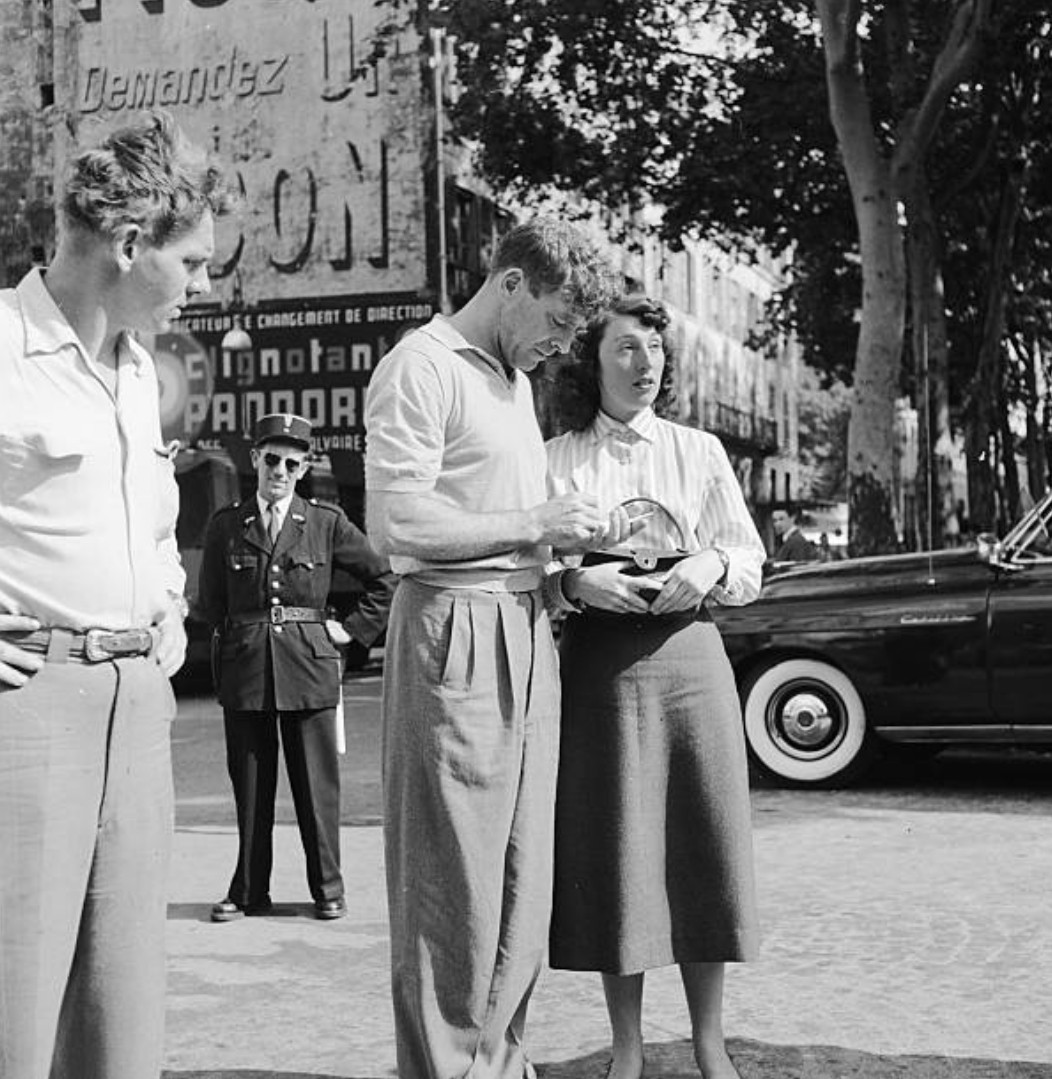 Some people standing around. Photo is from the mid-century. Man has a knitted polo and full legged, pleated pants. 