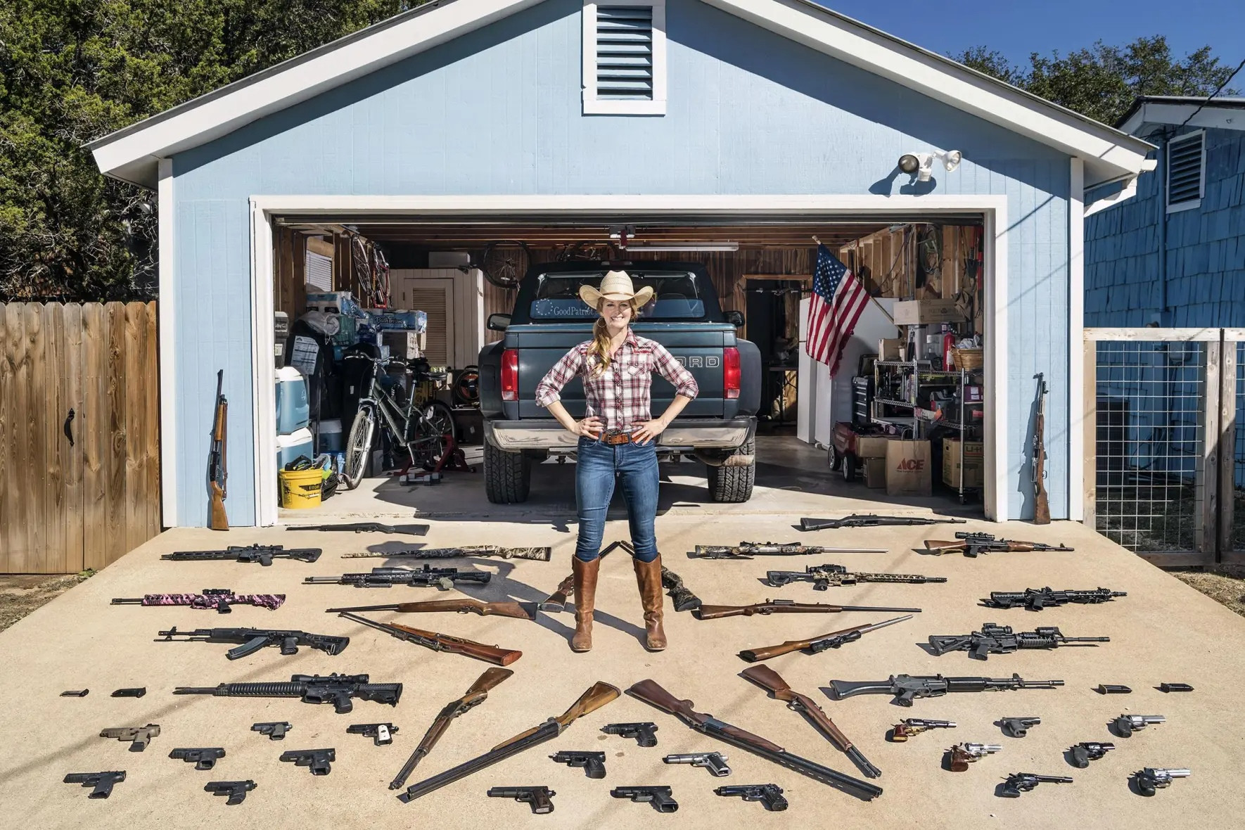A woman outside her garage. The front of her home is full of guns, some laid out in a star formation. 